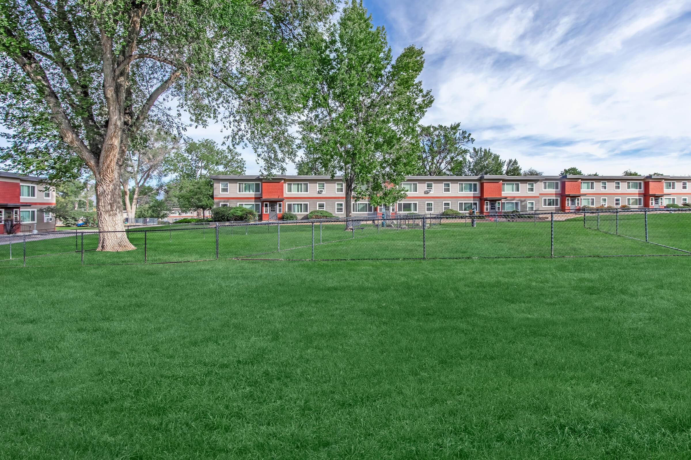 A green grassy field in front of a row of residential buildings, featuring trees and a fence. The buildings have a combination of red and gray siding, with clear blue skies and scattered clouds above. The scene suggests a well-maintained community environment.