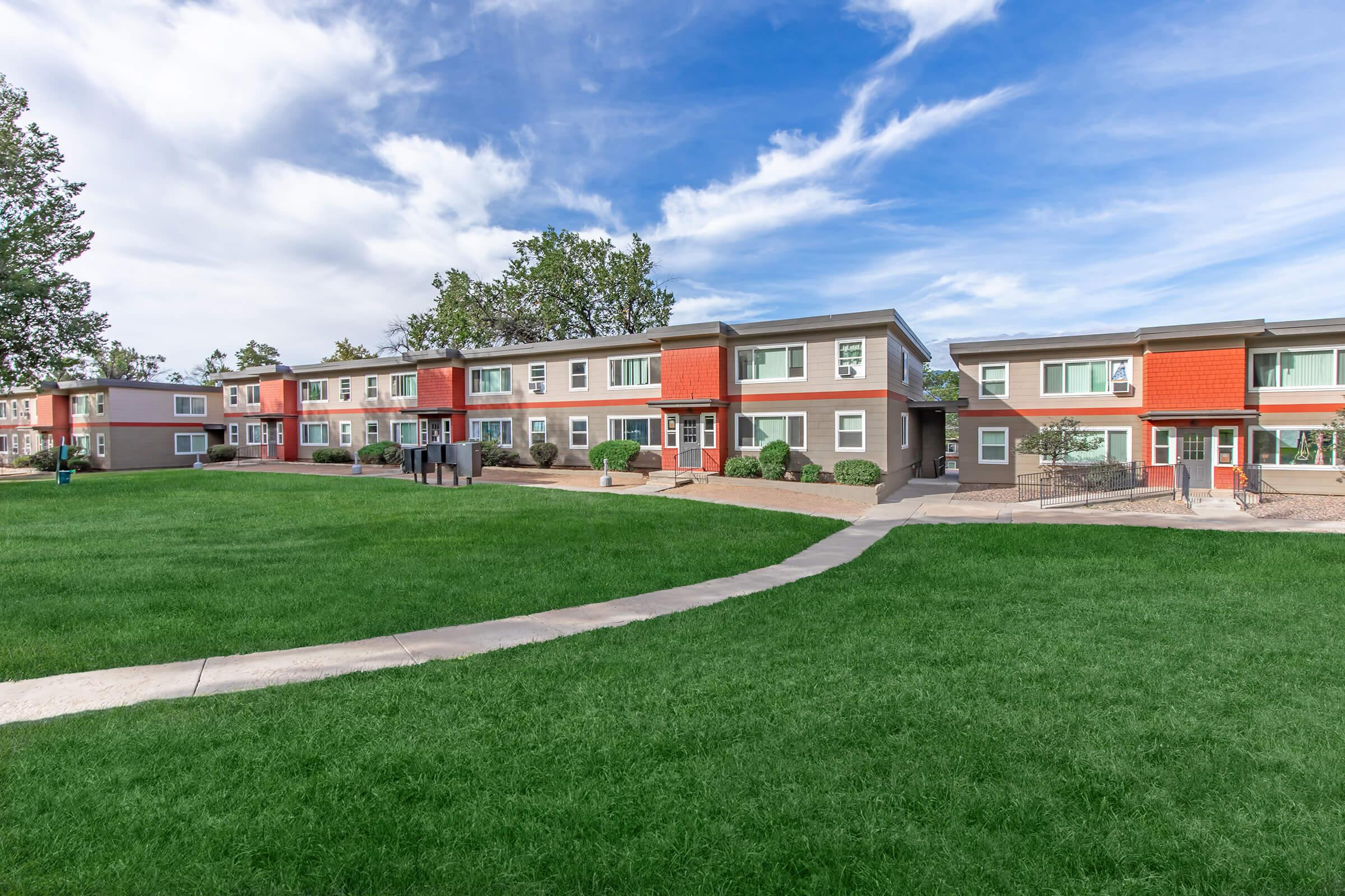A view of a modern apartment complex featuring multiple two-story buildings with red and gray exteriors. The complex is surrounded by well-maintained green lawns and pathways, under a clear blue sky with some clouds.