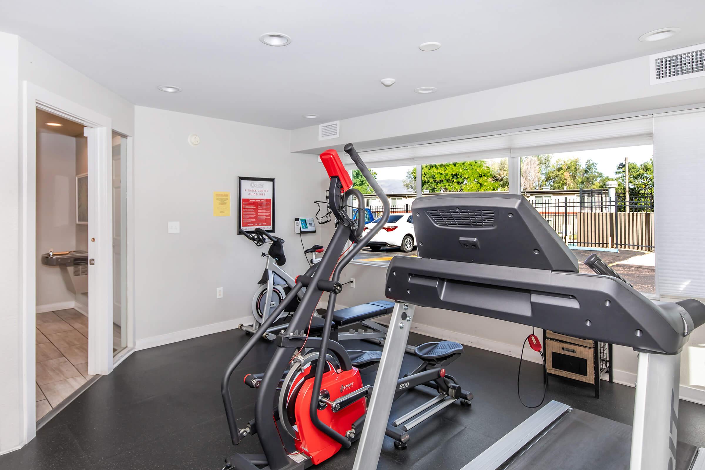A modern gym interior featuring a treadmill, stationary bike, and weight bench. Natural light enters through a large window, providing a view of the outside. The space is clean and well-lit, with fitness equipment positioned neatly and motivational signs on the walls.
