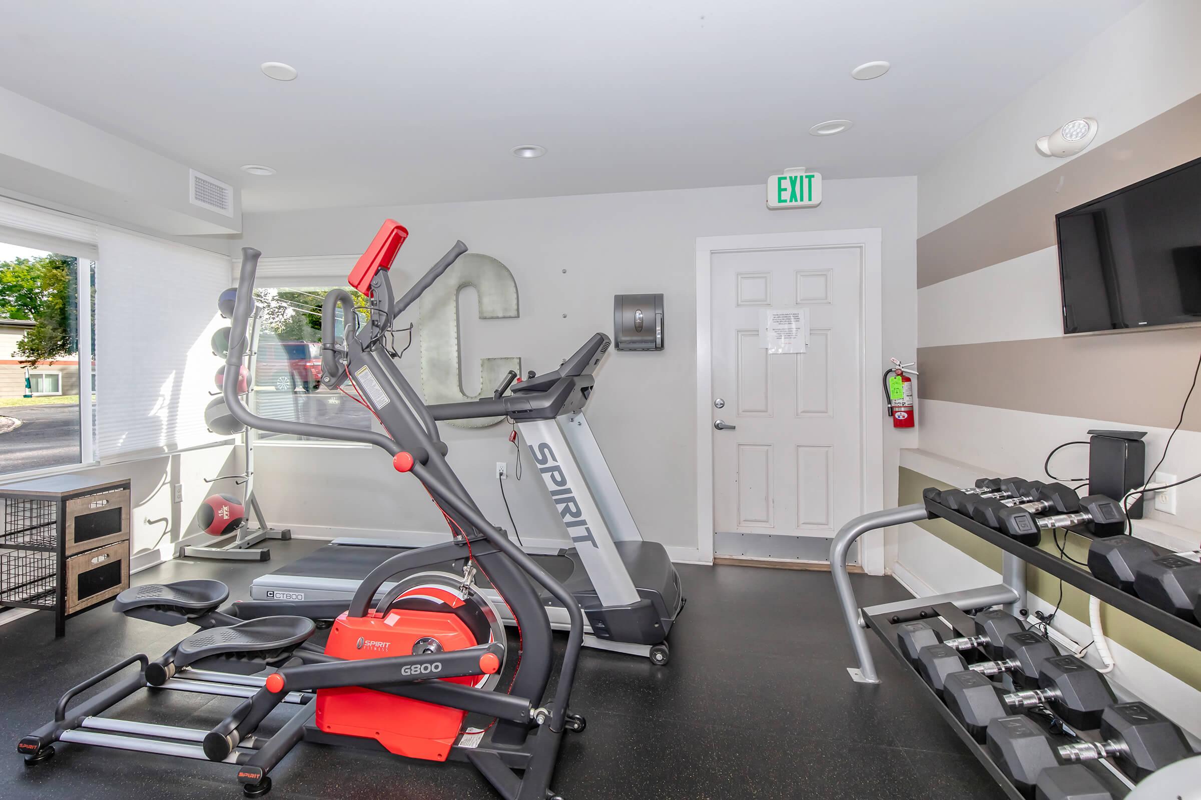 A modern gym interior featuring various exercise equipment, including an elliptical machine, a treadmill, and a weight rack with dumbbells. The space has large windows for natural light, a door leading outside, and a large wall-mounted television. The walls are decorated with horizontal stripes in neutral colors.