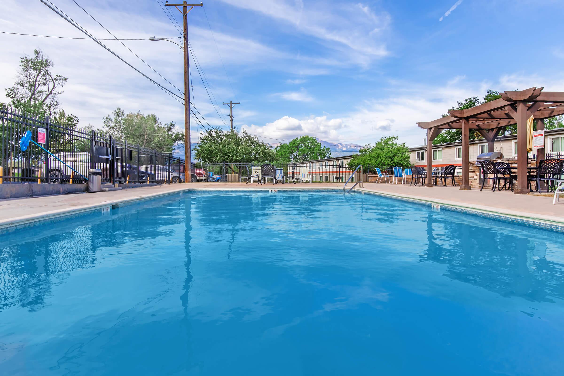 A clean, inviting swimming pool surrounded by a concrete deck, with lounge chairs and shaded seating under a wooden pergola. Trees and a cloudy blue sky are visible in the background, along with power lines and a glimpse of nearby buildings. The pool surface reflects the sky, creating a serene atmosphere.