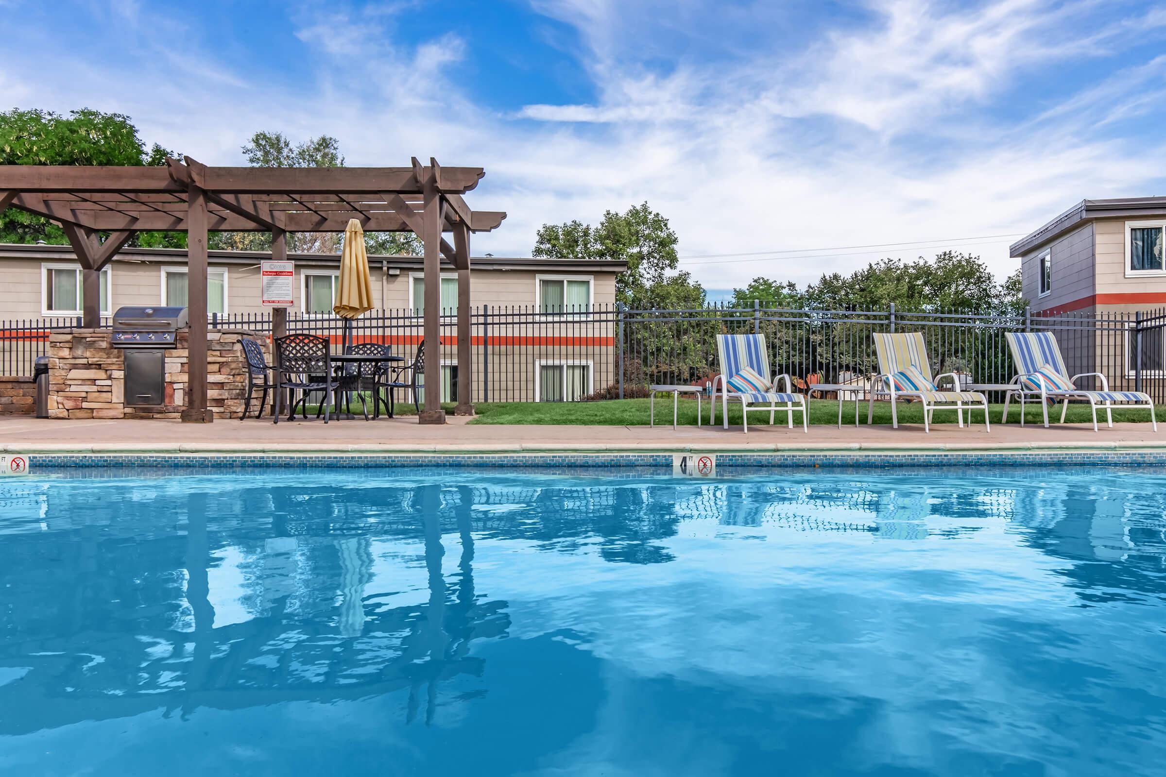 View of a swimming pool with clear blue water, featuring a pergola with outdoor furniture, including tables and chairs. In the background, there are residential buildings and a grassy area. The sky is partly cloudy, enhancing the relaxed outdoor atmosphere.