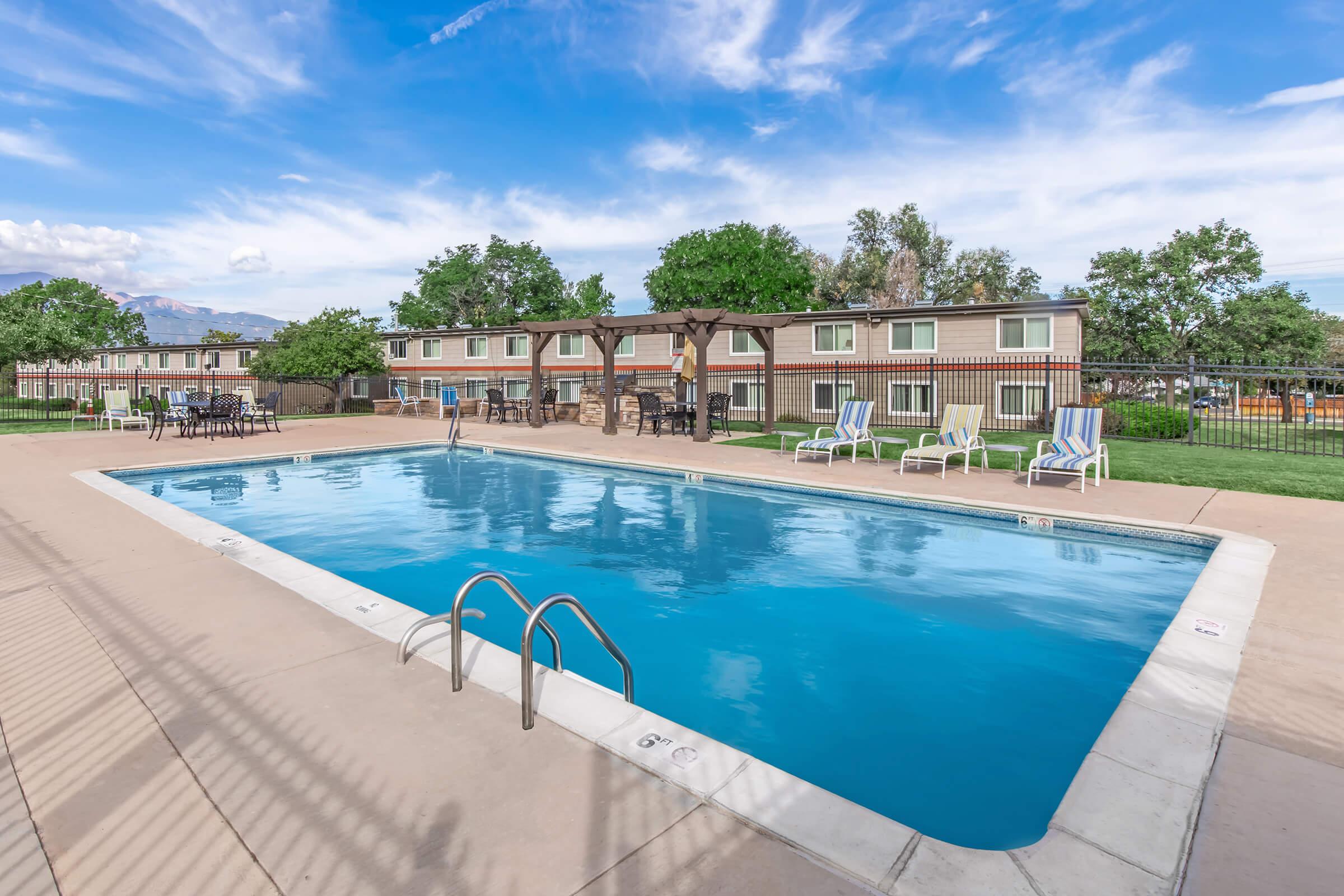 A clear blue swimming pool surrounded by lounge chairs, with a pergola structure nearby. The pool area is well-lit and set against a backdrop of green grass and trees, with a row of buildings visible in the background under a bright blue sky.