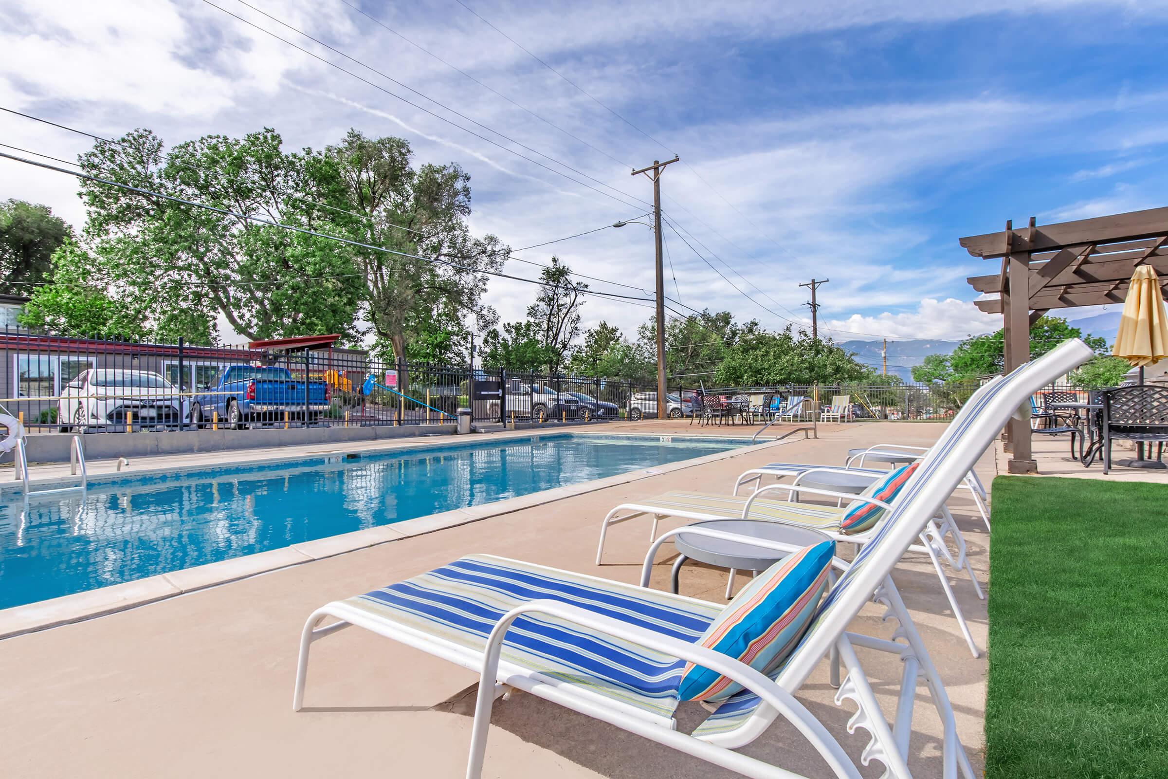 A brightly lit outdoor swimming pool area featuring a clear blue pool with lounge chairs on one side. In the background, there are trees, parked cars, and a pergola with seating. The sky is partly cloudy, creating a pleasant atmosphere for relaxation.