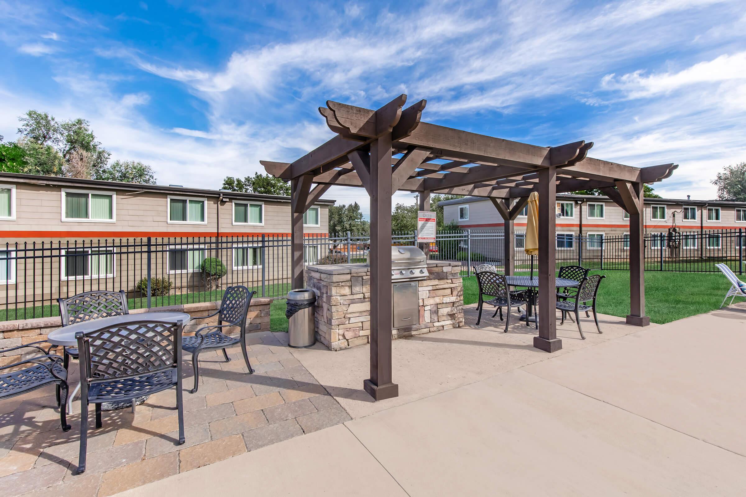 Outdoor grilling area under a wooden pergola, featuring a stone grill. Surrounding are metal tables and chairs, with green lawn in the background. Apartment buildings with windows are visible behind the grilling space. The sky is partly cloudy, creating a pleasant atmosphere.