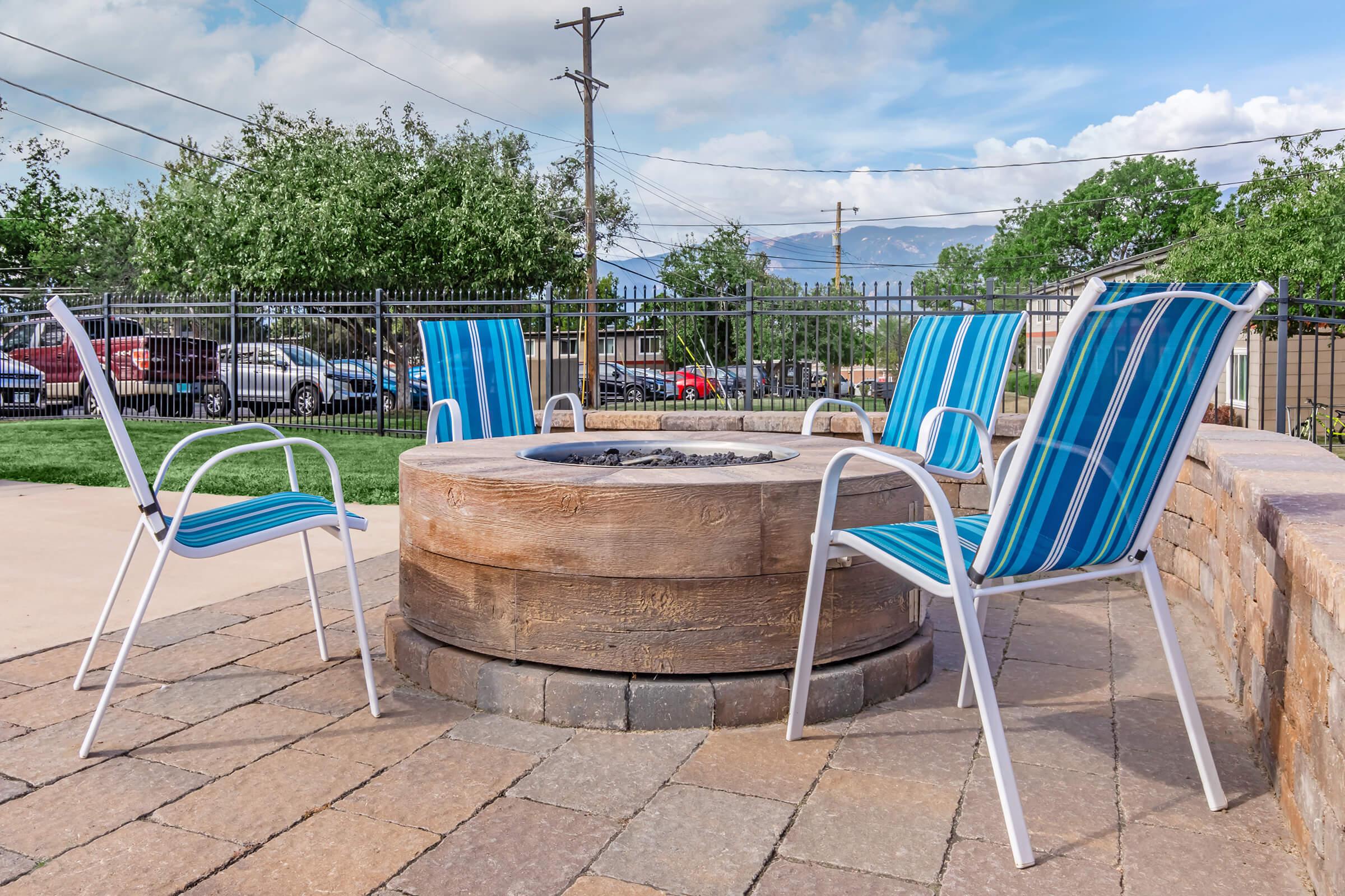 A circular fire pit made of stone, surrounded by four striped lounge chairs. The setting includes green grass and trees in the background, with a clear blue sky and distant mountains. Nearby parking is visible, suggesting a communal outdoor area.