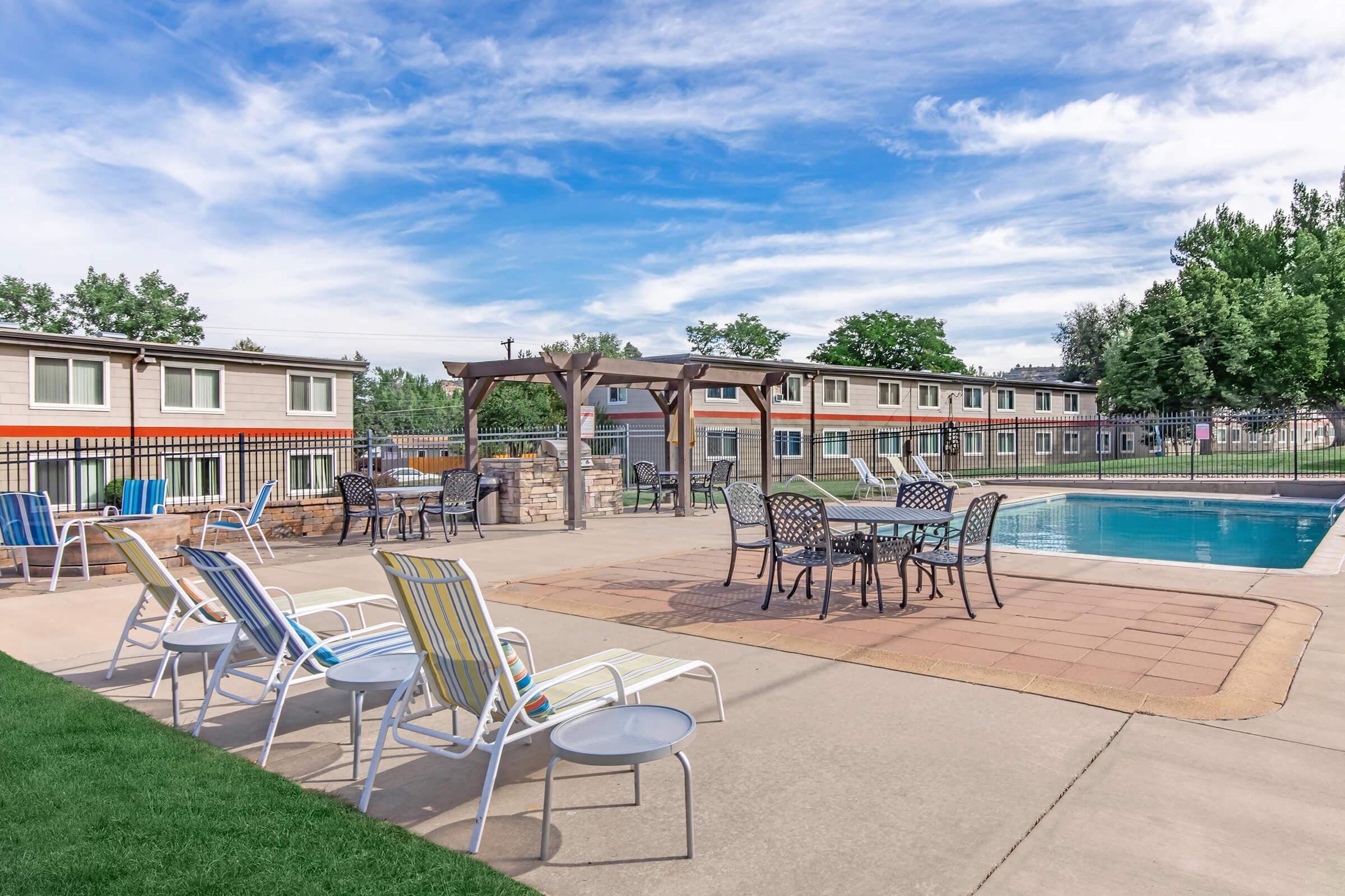 A sunny outdoor pool area featuring a clear blue pool surrounded by lounge chairs. There are tables with chairs for seating beneath a wooden pergola. In the background, multi-story buildings with green trees line the area, and the sky is clear with scattered clouds.