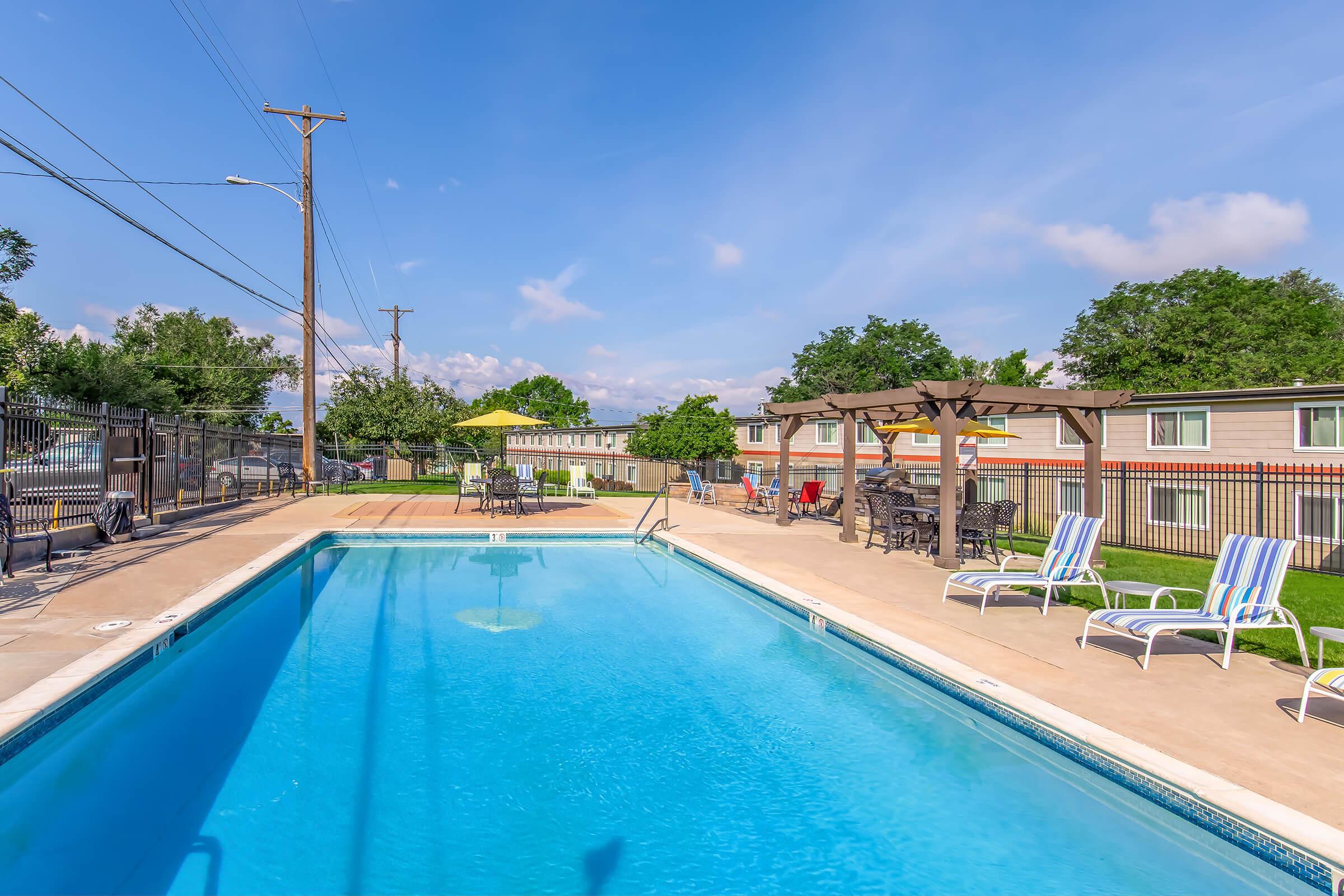 A clear blue swimming pool surrounded by a patio area with lounge chairs. There are green trees in the background, with a gazebo and tables for seating. The sky is bright with a few clouds, and there are power lines visible. The setting suggests a relaxed outdoor atmosphere suitable for leisure.
