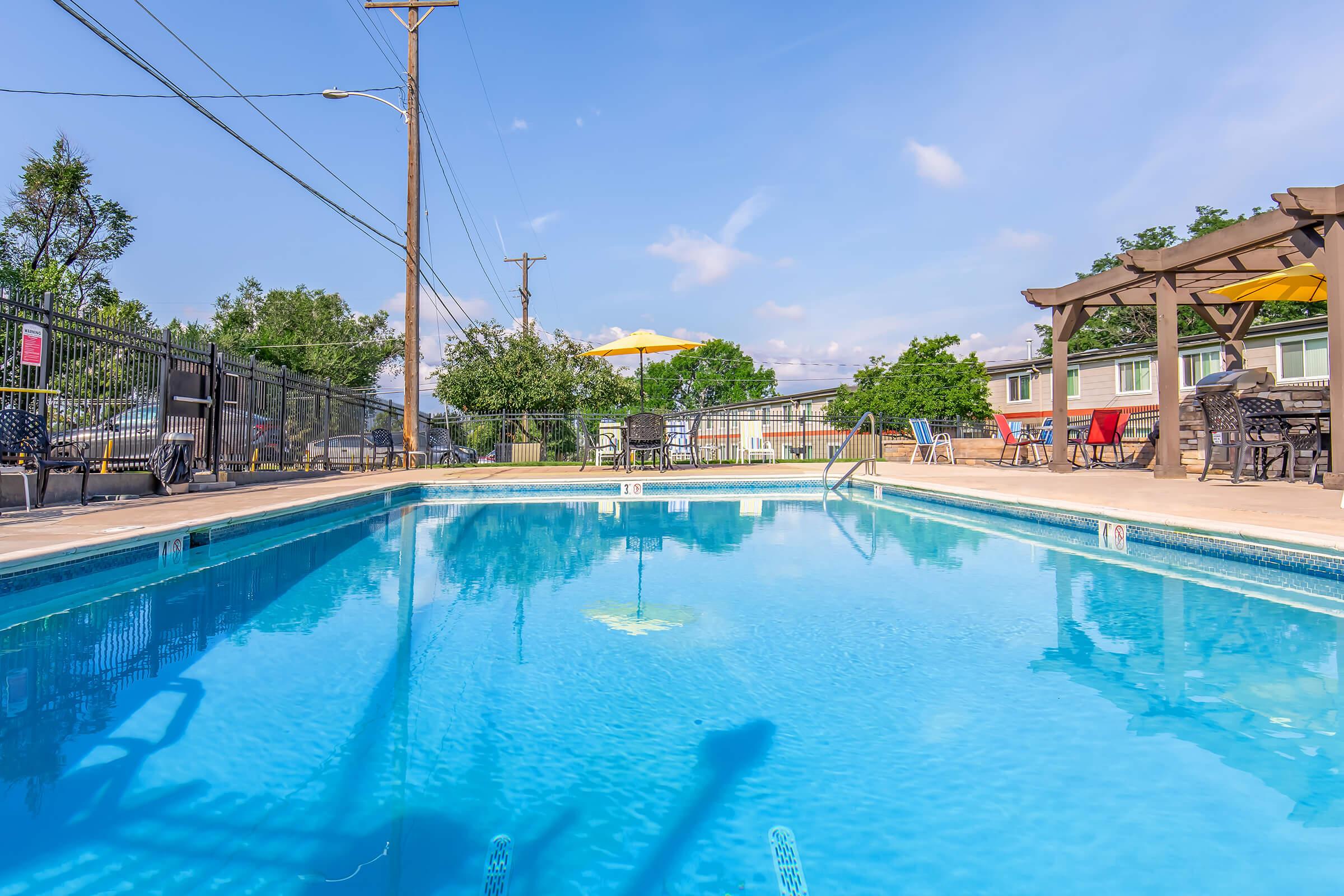 A clear blue swimming pool with a deck area, surrounded by lounge chairs and umbrellas. Trees and a building are visible in the background under a sunny sky. Utility poles are seen nearby, emphasizing the outdoor setting. The water appears inviting and clean.