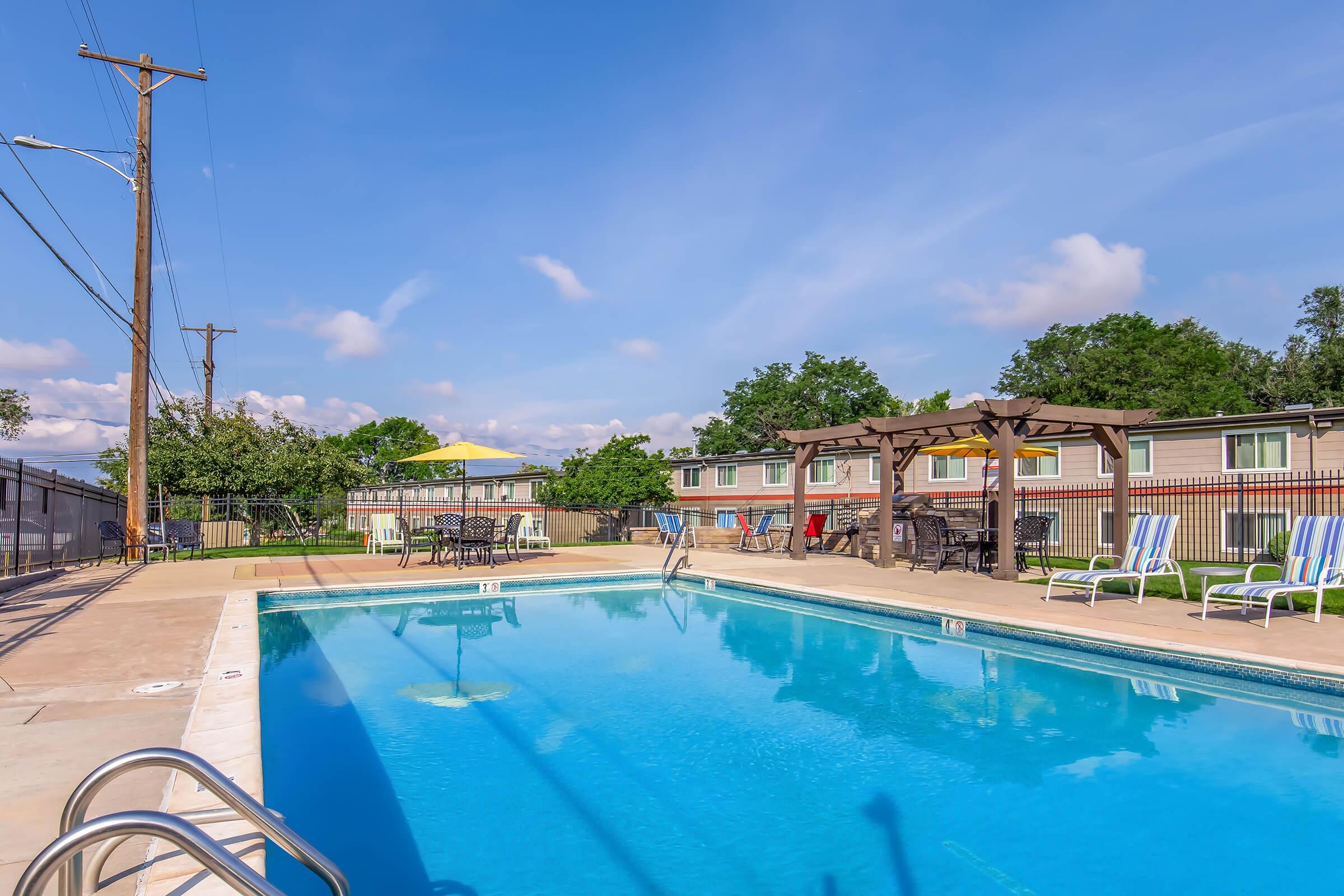 A clear blue swimming pool surrounded by lounge chairs and tables, with green trees and a few buildings in the background. The sky is bright with fluffy clouds, and there's a pergola near the pool area. Power lines are visible along the edge of the scene.