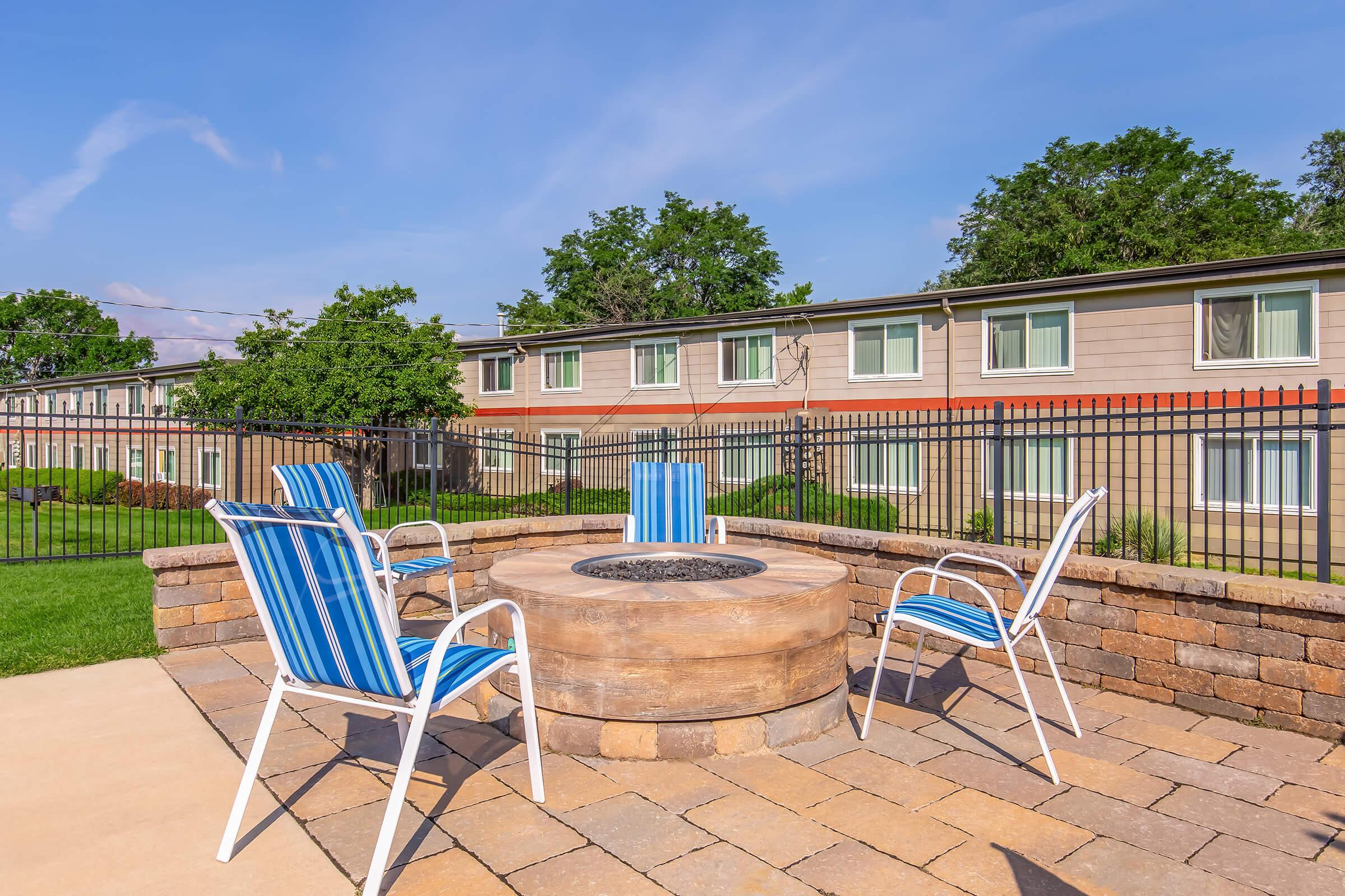 A cozy outdoor seating area featuring four striped blue and white chairs arranged around a circular stone fire pit. In the background, there are buildings with several windows and greenery, under a clear blue sky. The space is enclosed with a black fence, creating a welcoming atmosphere for relaxation.