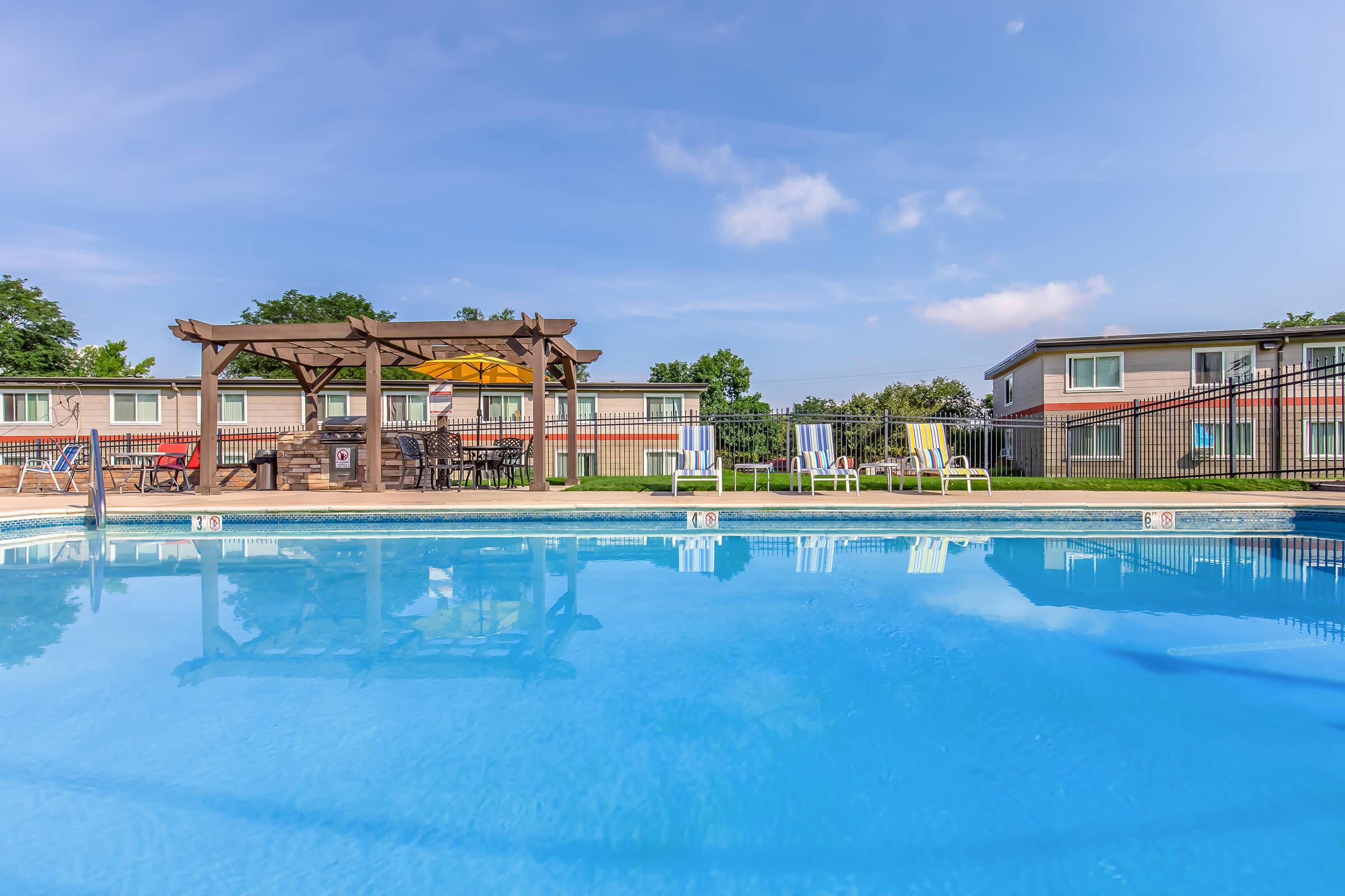 A clear blue swimming pool surrounded by lounge chairs and a shaded seating area under a pergola. In the background, there are two-story buildings with a lawn area. The sky is bright with a few clouds, creating a pleasant atmosphere for relaxation and leisure.