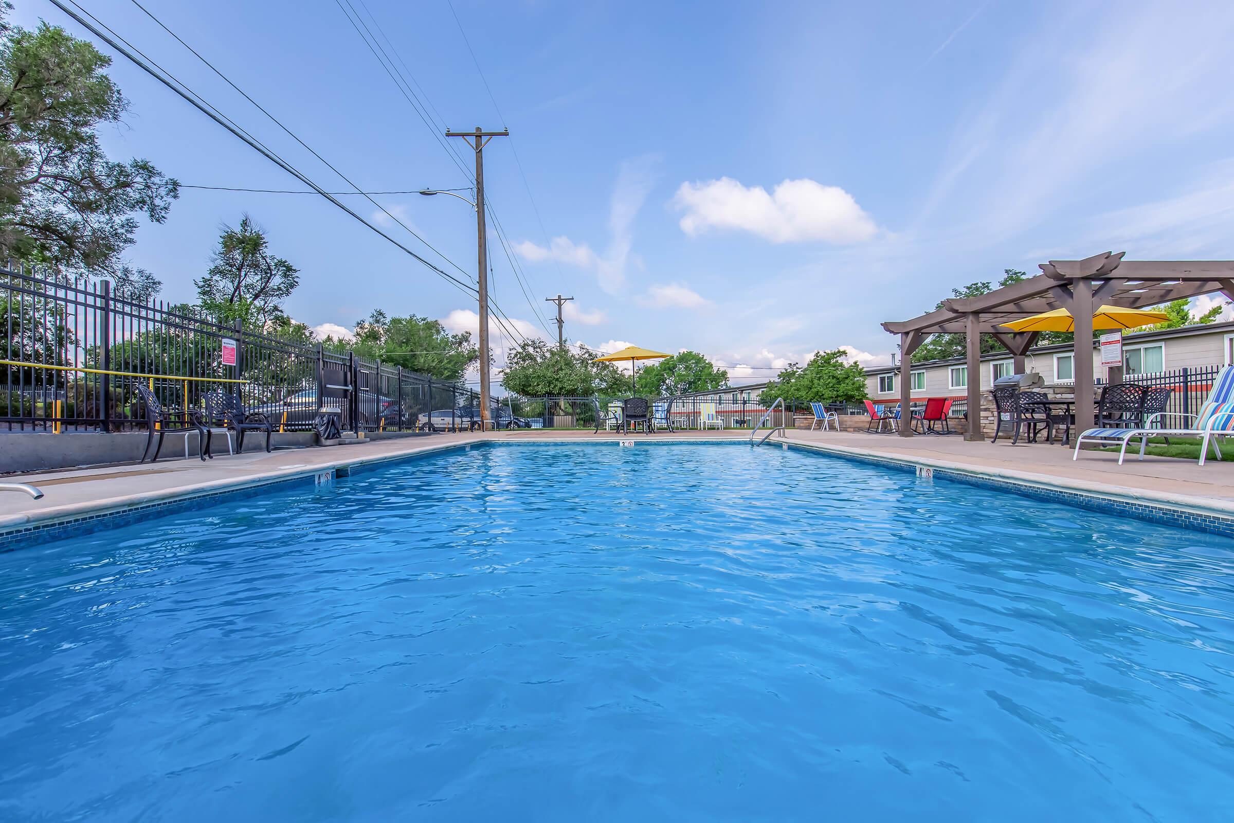 A clear blue swimming pool surrounded by lounge chairs and umbrellas, with trees and buildings in the background. The sky is partly cloudy, and there are power lines visible nearby. The scene conveys a relaxing outdoor atmosphere perfect for leisure activities.