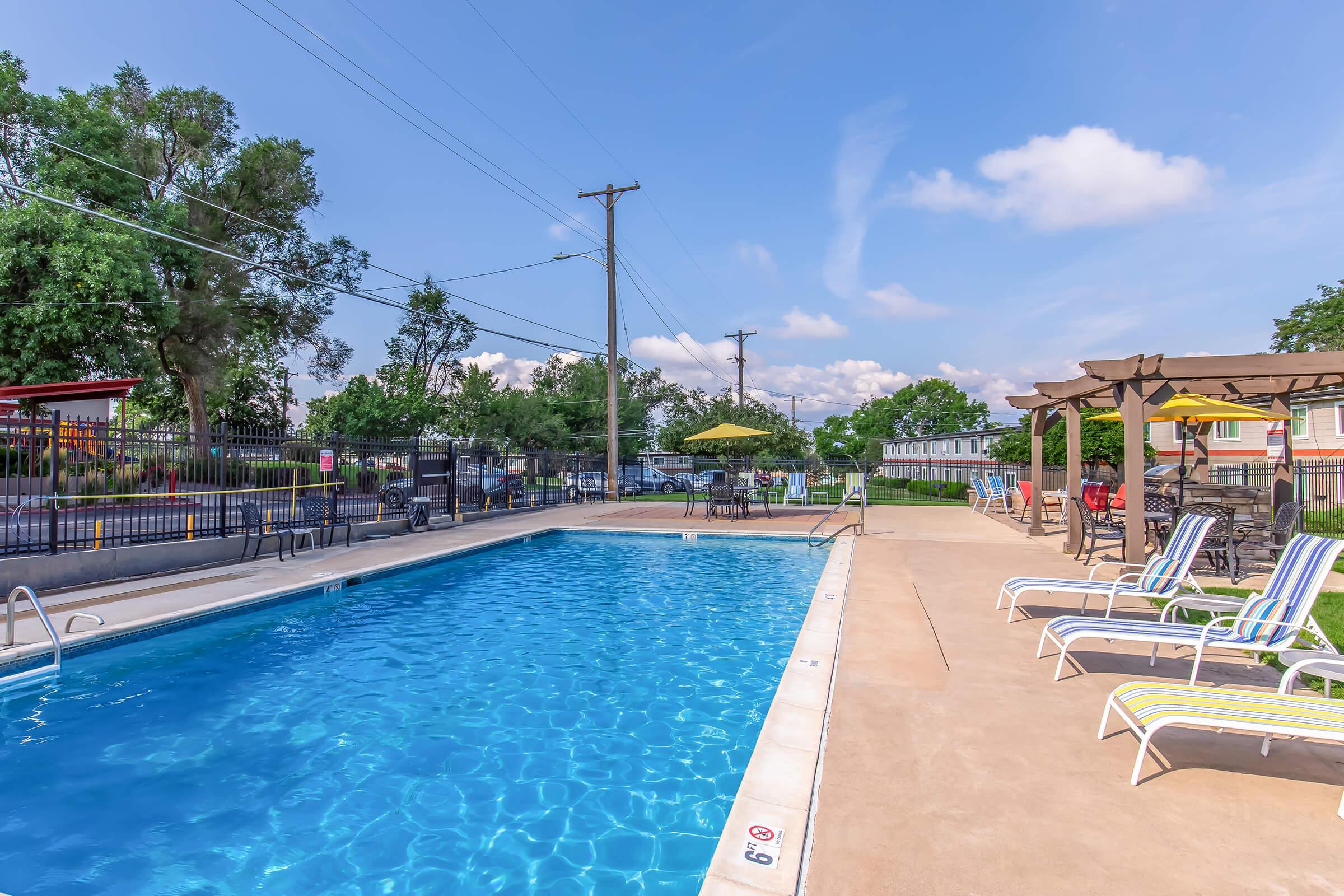 A clear swimming pool surrounded by lounge chairs and umbrellas on a sunny day. Trees and a fence line the background, with utility poles visible. The area is well-maintained and inviting, perfect for relaxation and leisure activities.