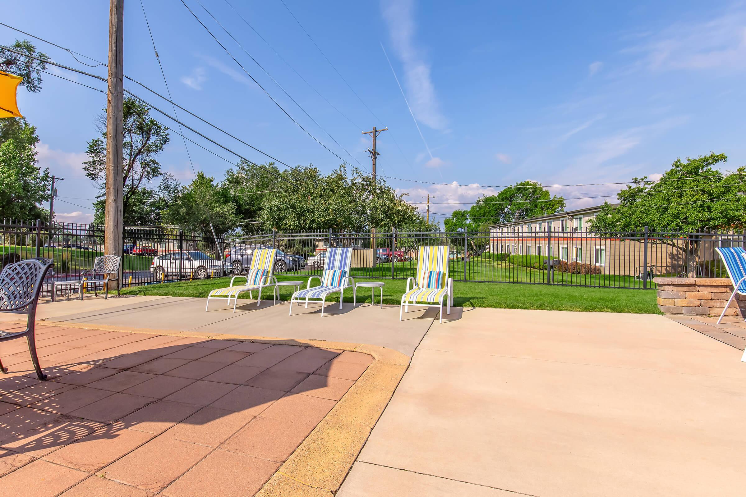 A sunny outdoor area featuring several striped lounge chairs arranged by a poolside. There is a grassy lawn in the background, along with a fence and trees. Power lines stretch across the sky, and a building can be seen in the distance, creating a relaxed atmosphere.