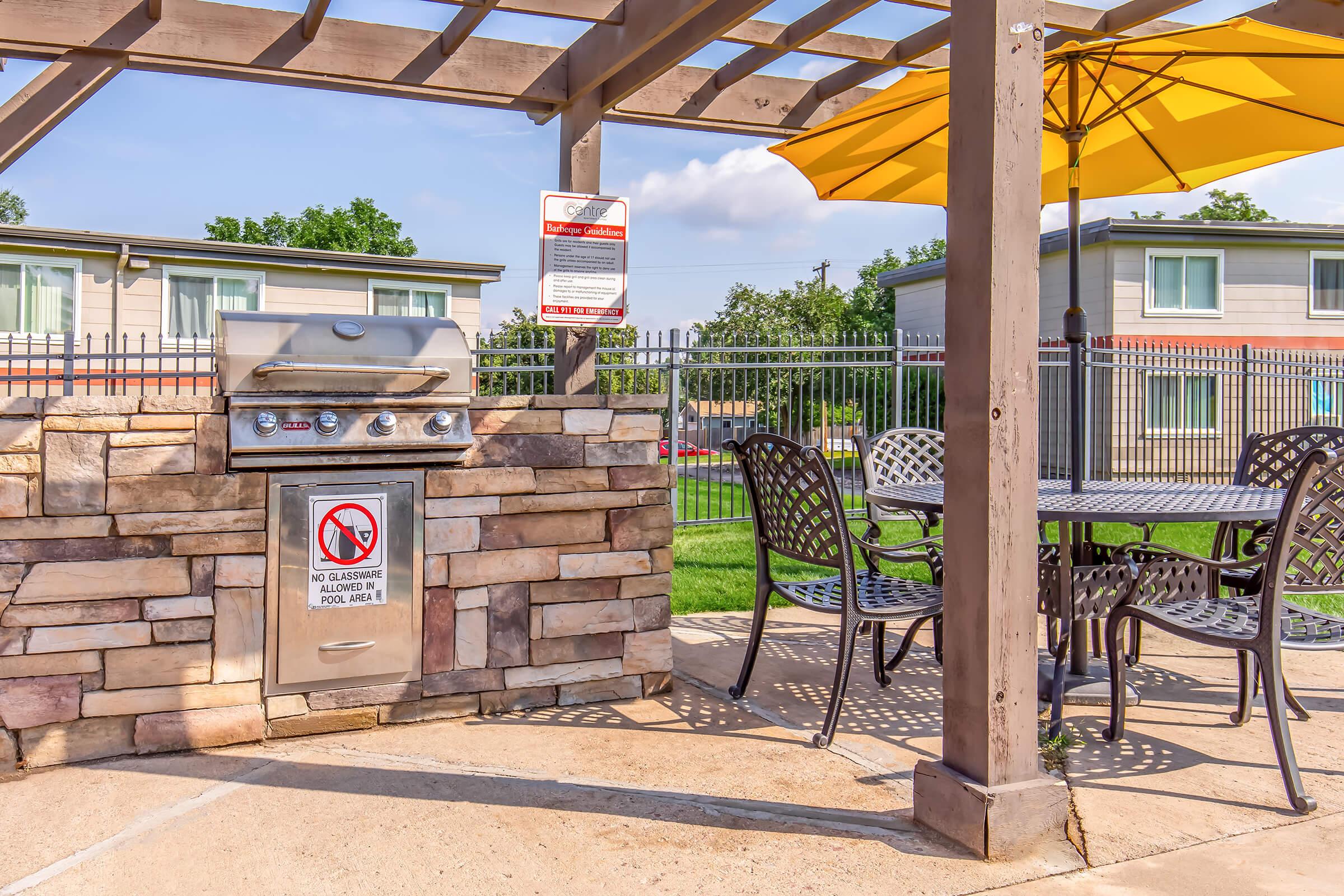 Outdoor grilling area featuring a stone BBQ grill under a shaded pergola. Nearby, a table and chairs are set up, complemented by a large yellow umbrella. A sign warns against swimming and grilling rules. In the background, a green lawn and apartments are visible.