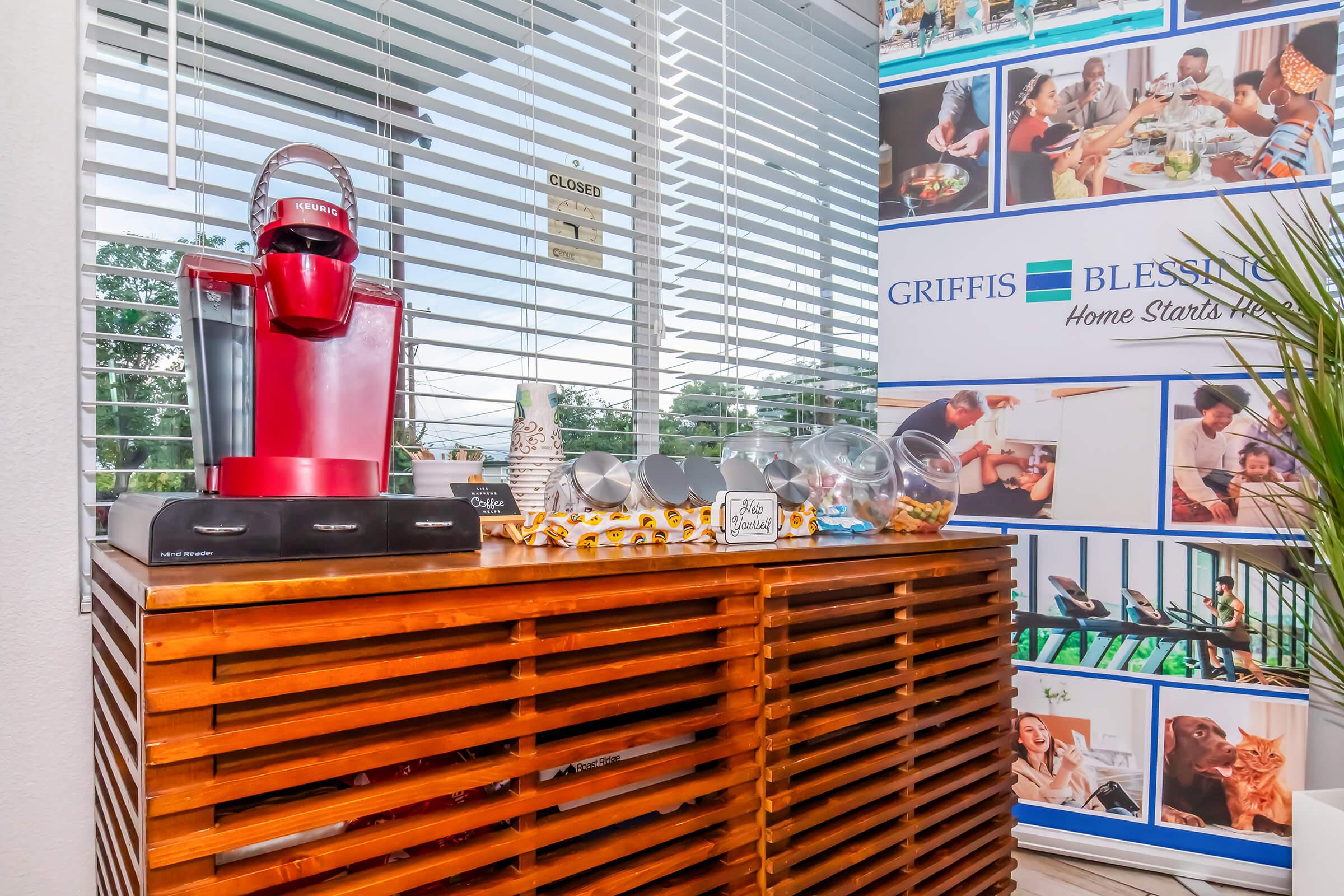A cozy corner featuring a red coffee maker on a wooden side table, decorated with a bowl of snacks. Behind it, a wall banner displays various images of people and activities, emphasizing a welcoming environment. Natural light streams in through the window, highlighting the inviting space.