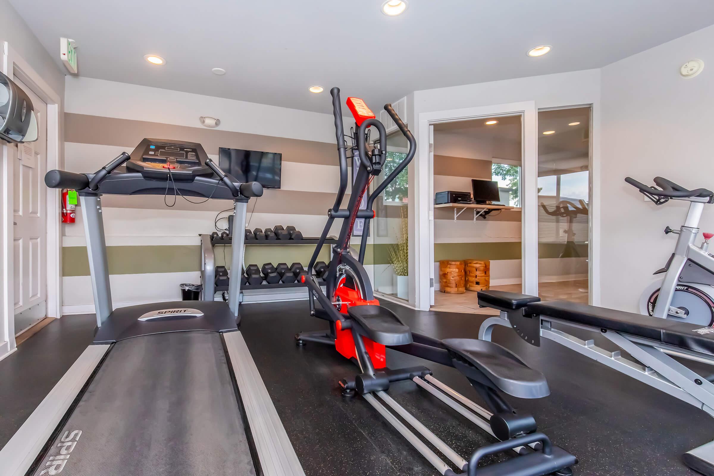 A modern gym interior featuring a treadmill, an elliptical machine, and a weight bench. In the background, there are free weights, a wall-mounted TV, and a workspace with a computer. The walls are painted in neutral tones, and ample lighting enhances the space.