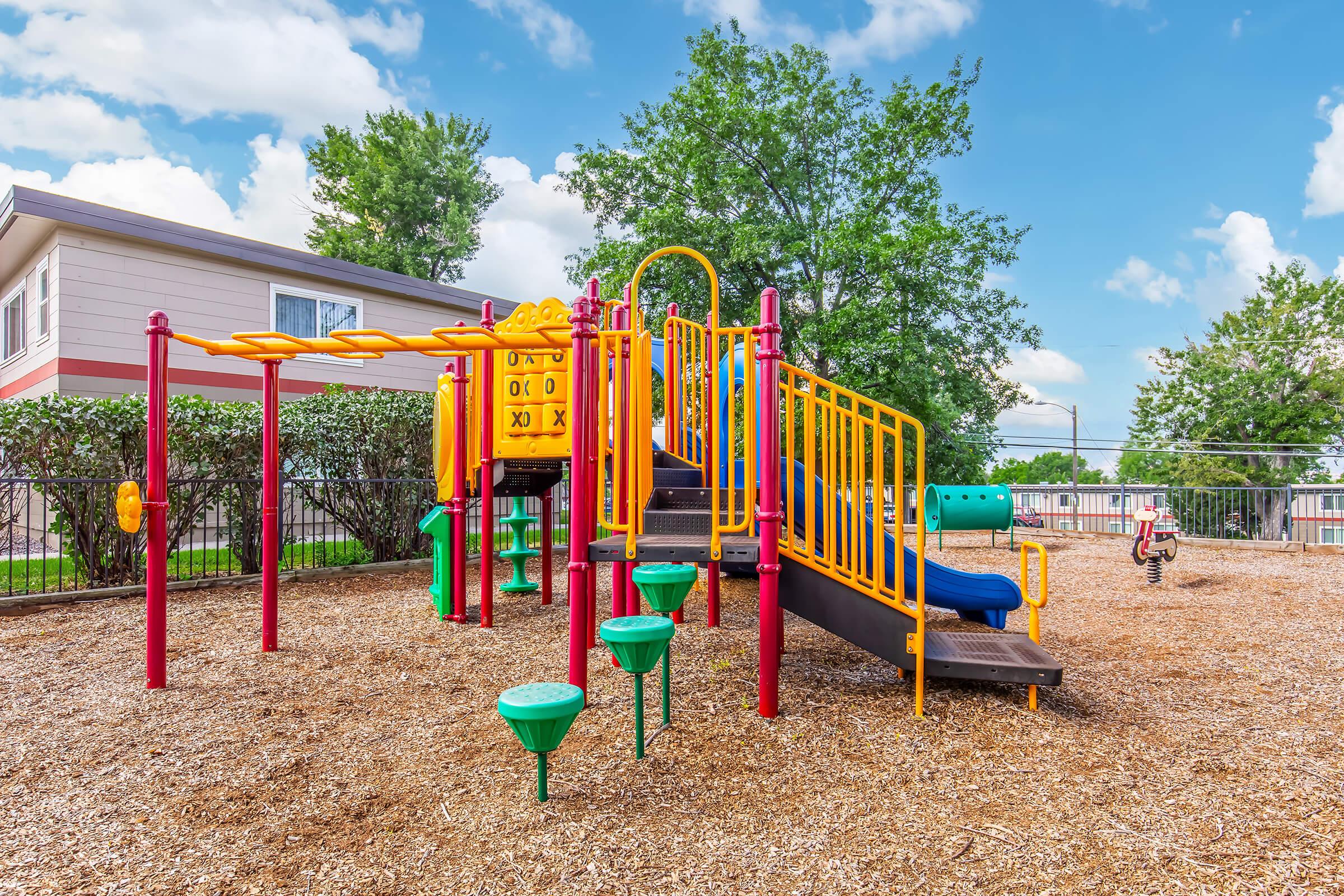 A colorful playground featuring a slide, climbing structure, and activity panels, surrounded by grass and trees. The area has a soft wood chip surface and is designed for children's play, providing a cheerful and inviting environment on a sunny day.