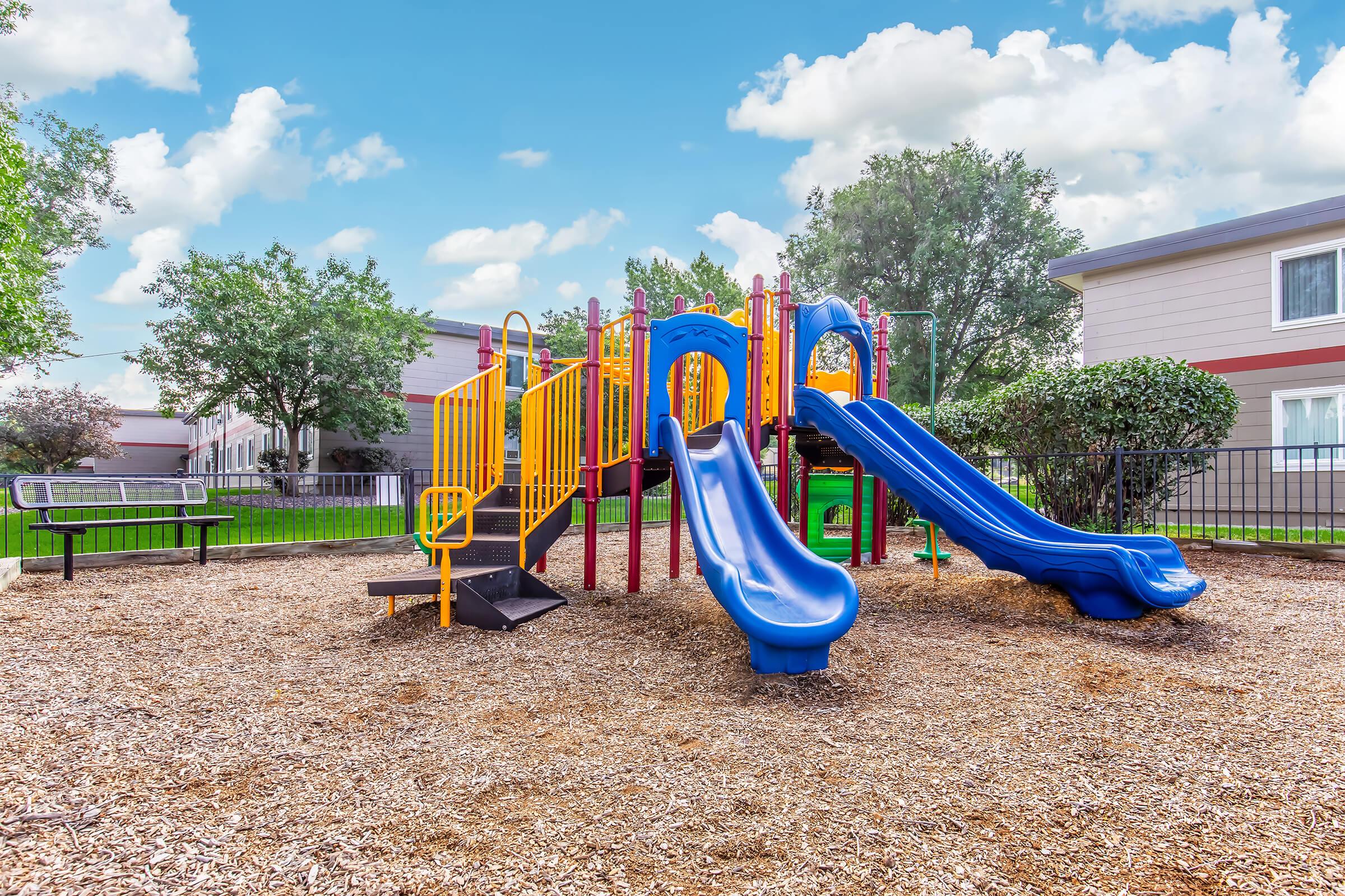 A colorful playground featuring two blue slides, climbing structures, and safety barriers, surrounded by grassy areas and trees. The scene is set under a bright blue sky with fluffy clouds, creating a cheerful atmosphere ideal for outdoor play.