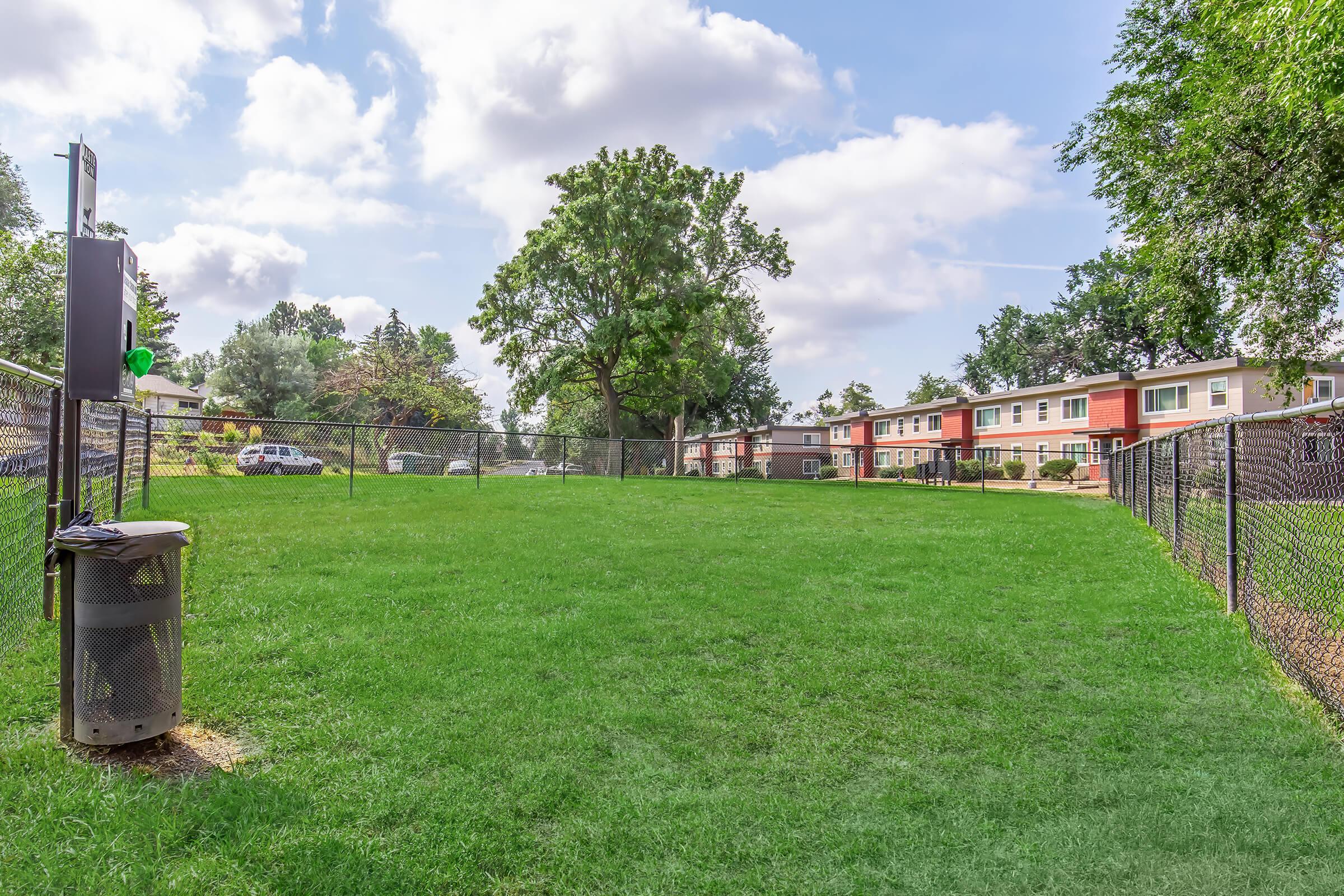 A green grassy area enclosed by a chain-link fence, with a trash can on the left. In the background, there are several two-story residential buildings surrounded by trees and bright blue skies with scattered clouds.