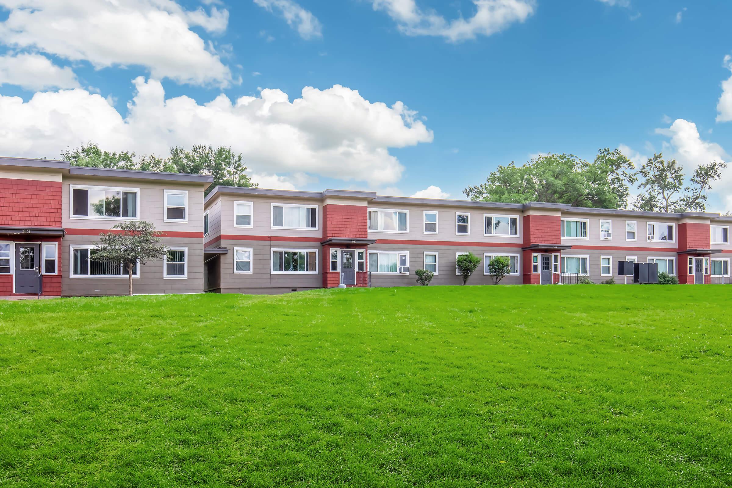 A contemporary two-story apartment complex is set against a bright blue sky with fluffy white clouds. The building features a mix of brown and red siding, with multiple windows and green lawns surrounding the structure. Trees are visible in the background, creating a welcoming atmosphere.