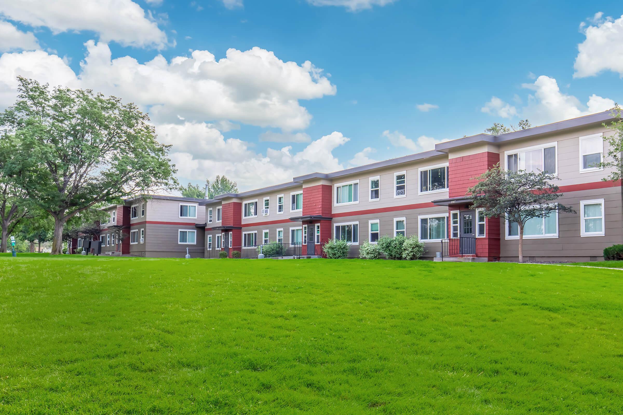 A modern apartment complex with multiple two-story buildings featuring a mix of red and gray exteriors. Lush green grass surrounds the buildings, and there are trees providing shade. The sky is bright blue with fluffy white clouds.
