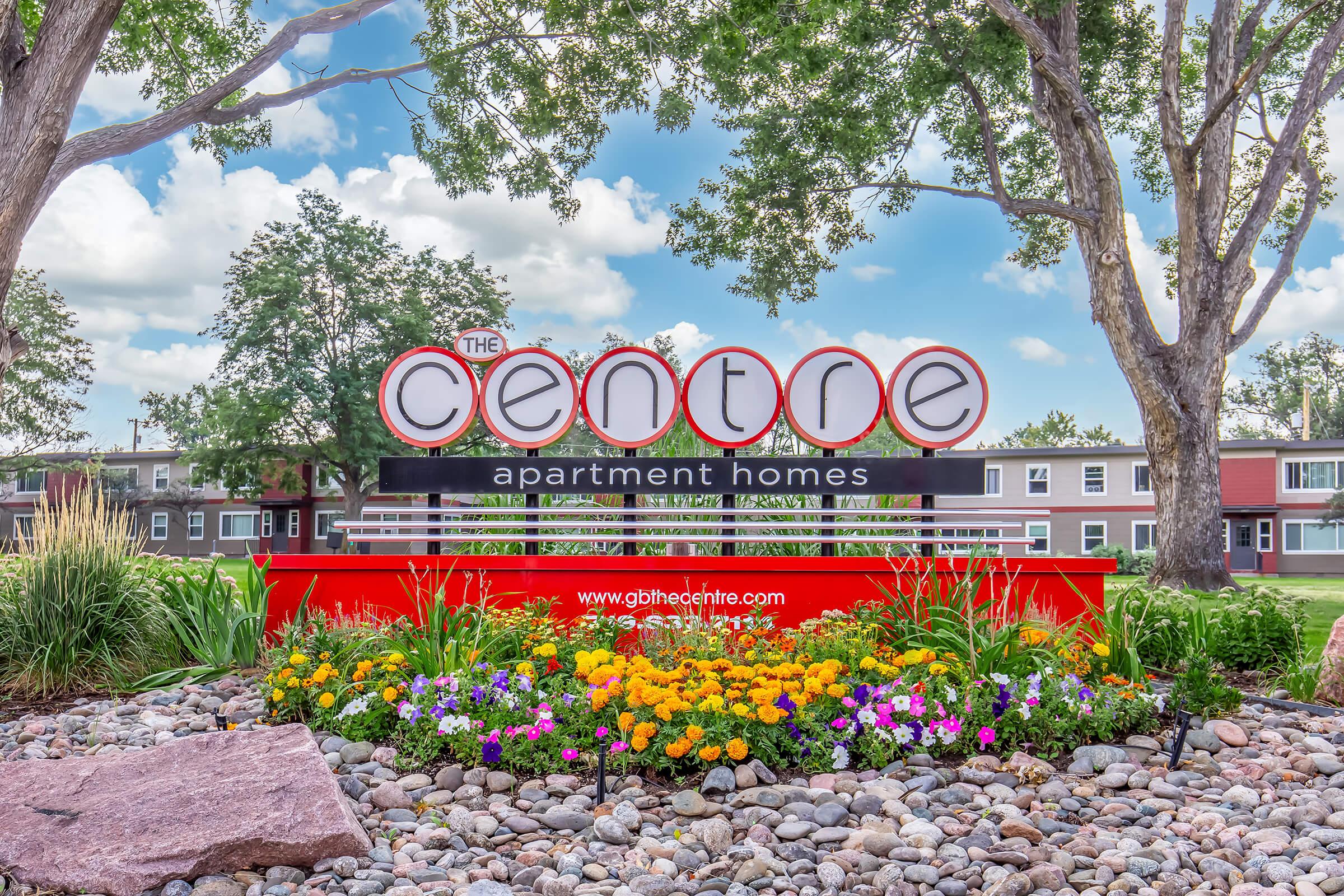 Large red and white sign for "The Centre" apartment homes, featuring the text prominently displayed. It is surrounded by colorful flower beds with various plants, rocks, and trees in the background under a partly cloudy sky. The website URL is visible at the bottom of the sign.