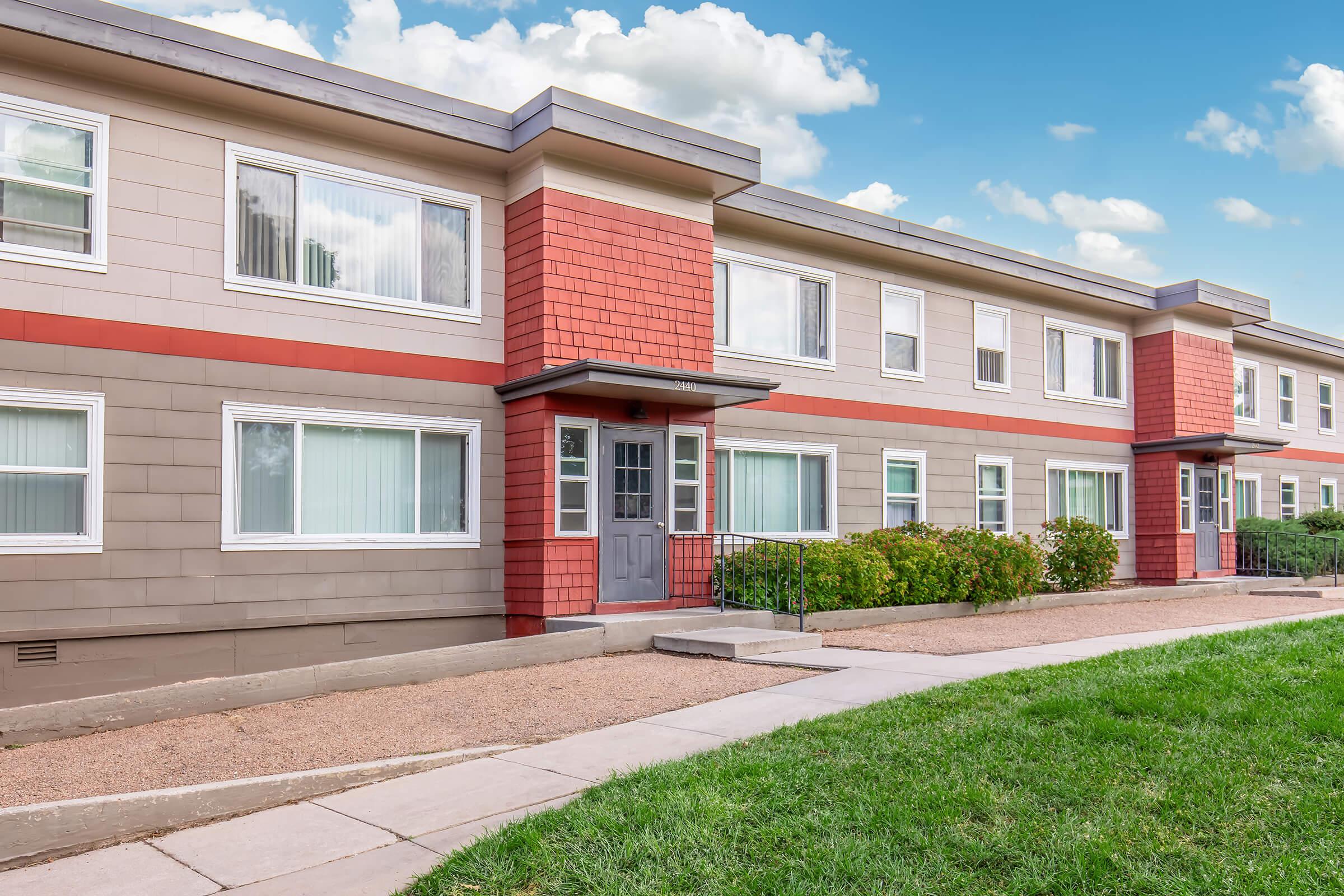 A modern apartment building with a mix of gray and red siding. The structure features multiple windows, a front door with steps leading up, and well-maintained landscaping. Green grass and a clear blue sky with clouds are visible in the background.