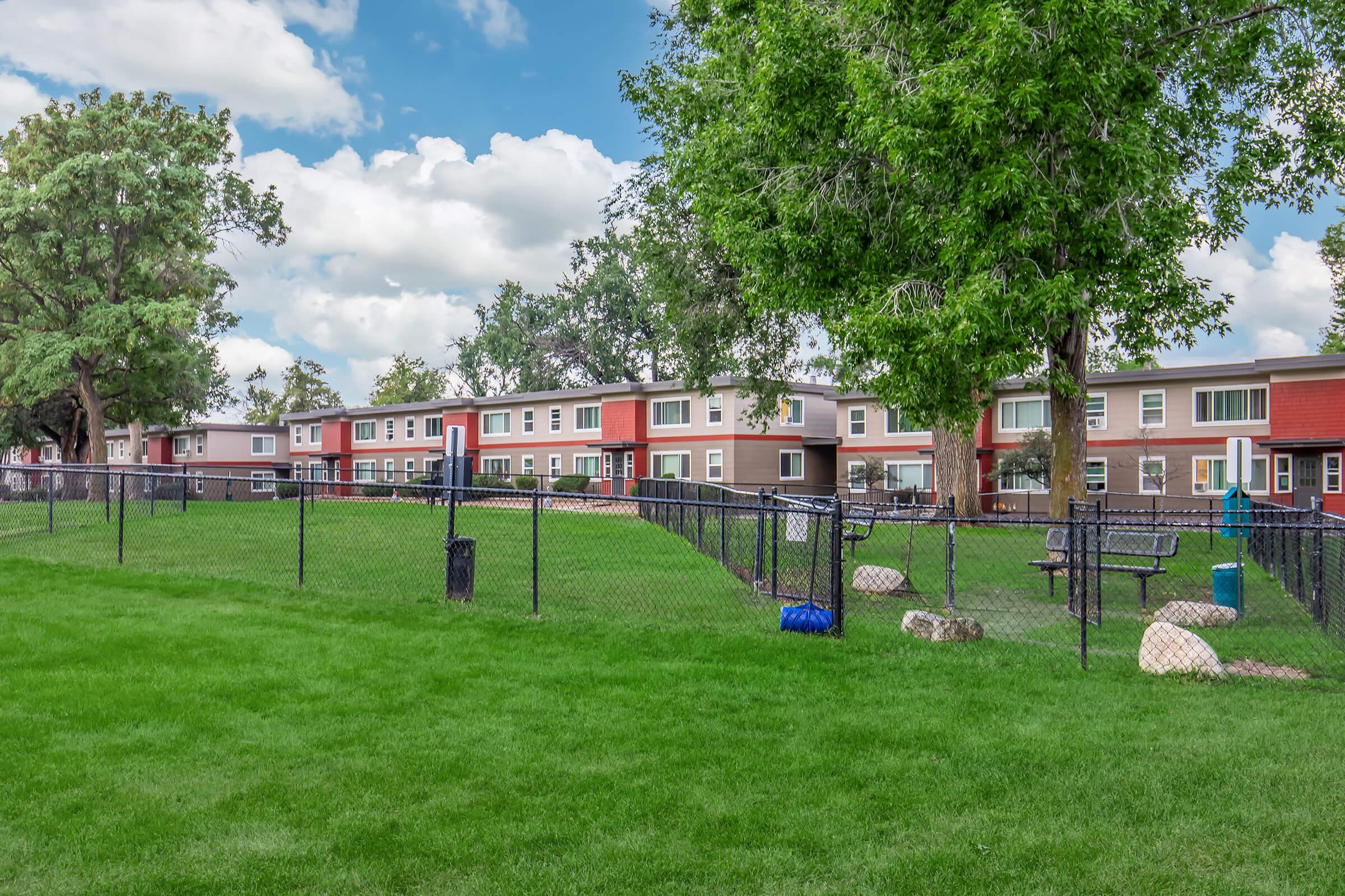 A grassy area enclosed by a black fence, featuring several residential buildings with red and neutral-colored exteriors. Trees provide shade, and there are benches and decorative boulders scattered around the lawn. The sky is partly cloudy, creating a pleasant outdoor atmosphere.