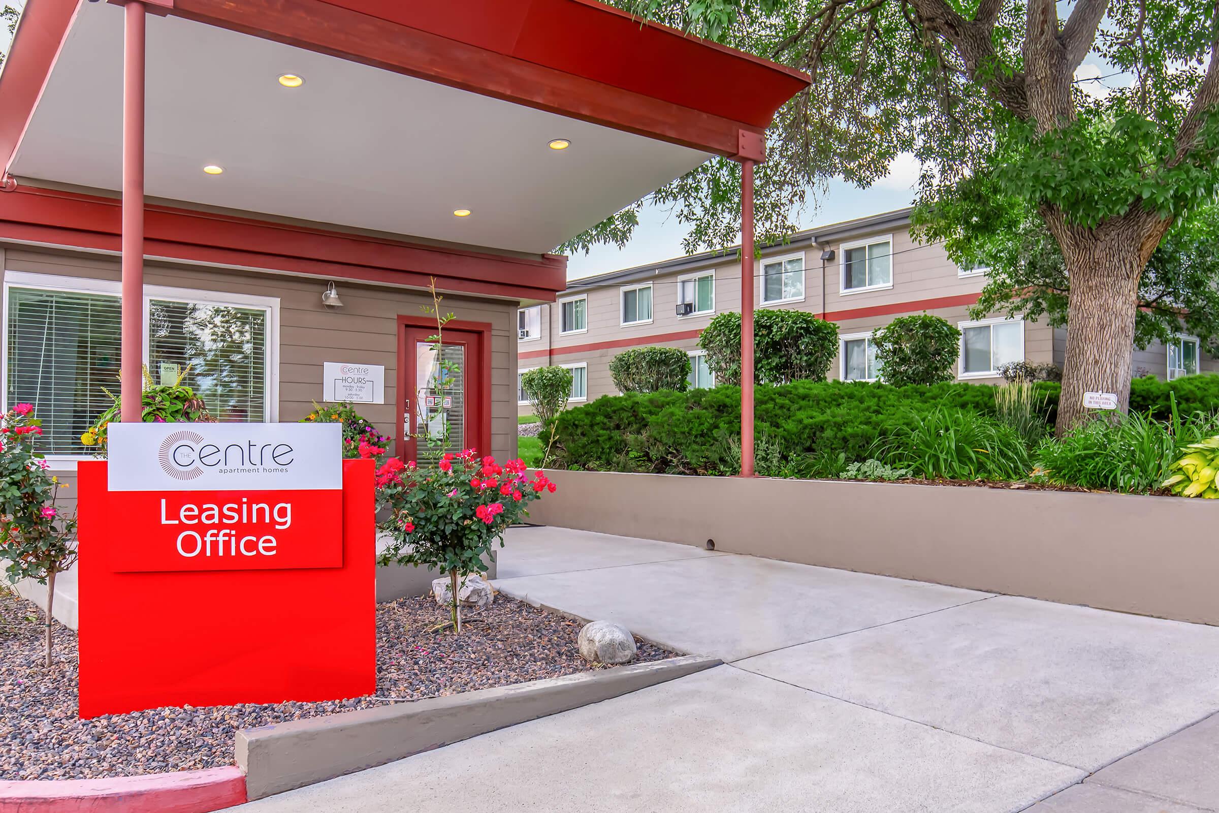 Leasing office exterior with a bright red sign reading "Centre Leasing Office." The entrance features a covered area with potted plants and a landscaped pathway. In the background, there are multiple buildings with windows and greenery, creating a welcoming environment.