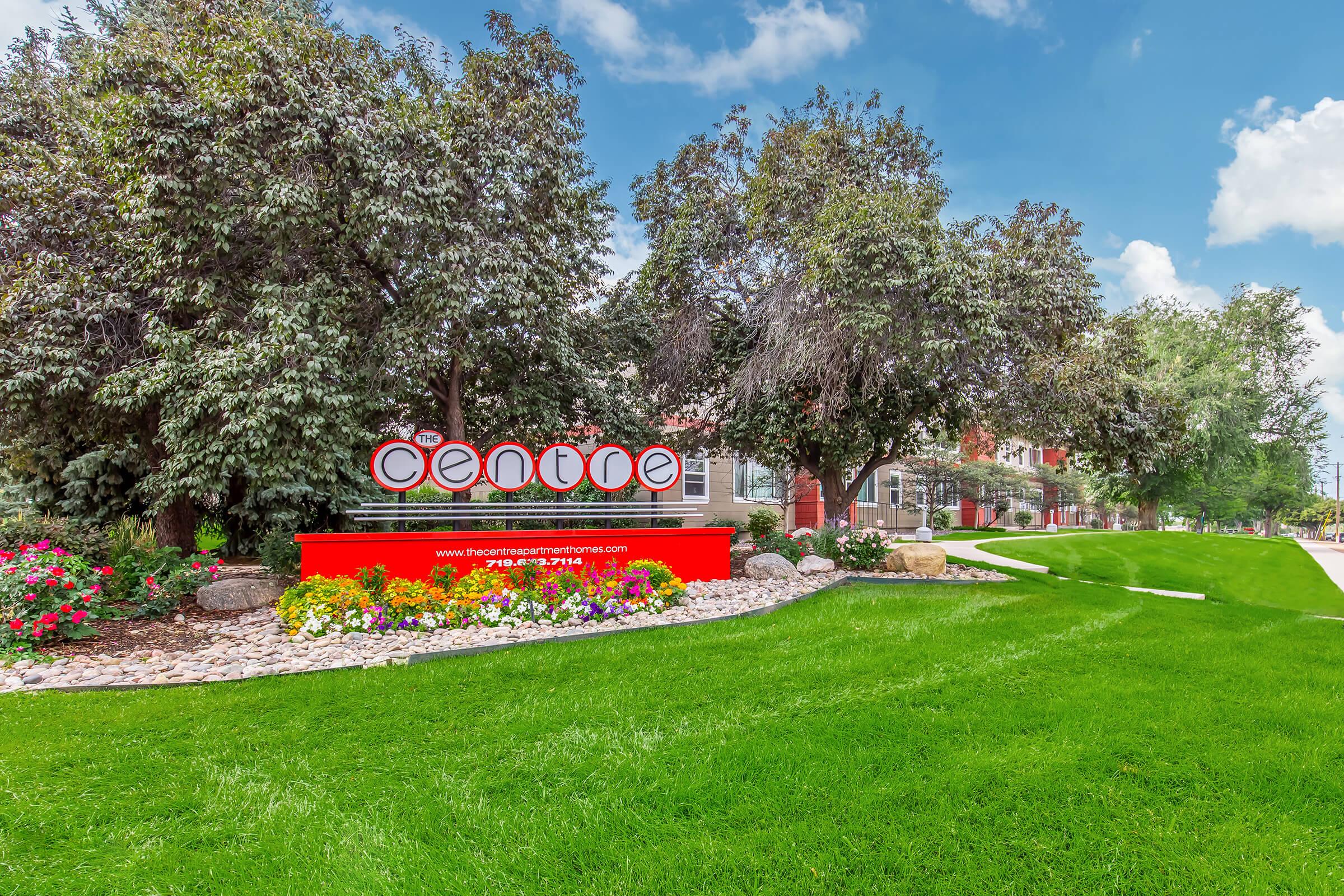 Sign featuring the word "centre" in bold letters, surrounded by colorful flower beds and greenery. A well-maintained lawn is visible, along with trees in the background and a blue sky with scattered clouds. The sign also includes contact information and an address, indicating a residential or commercial establishment.