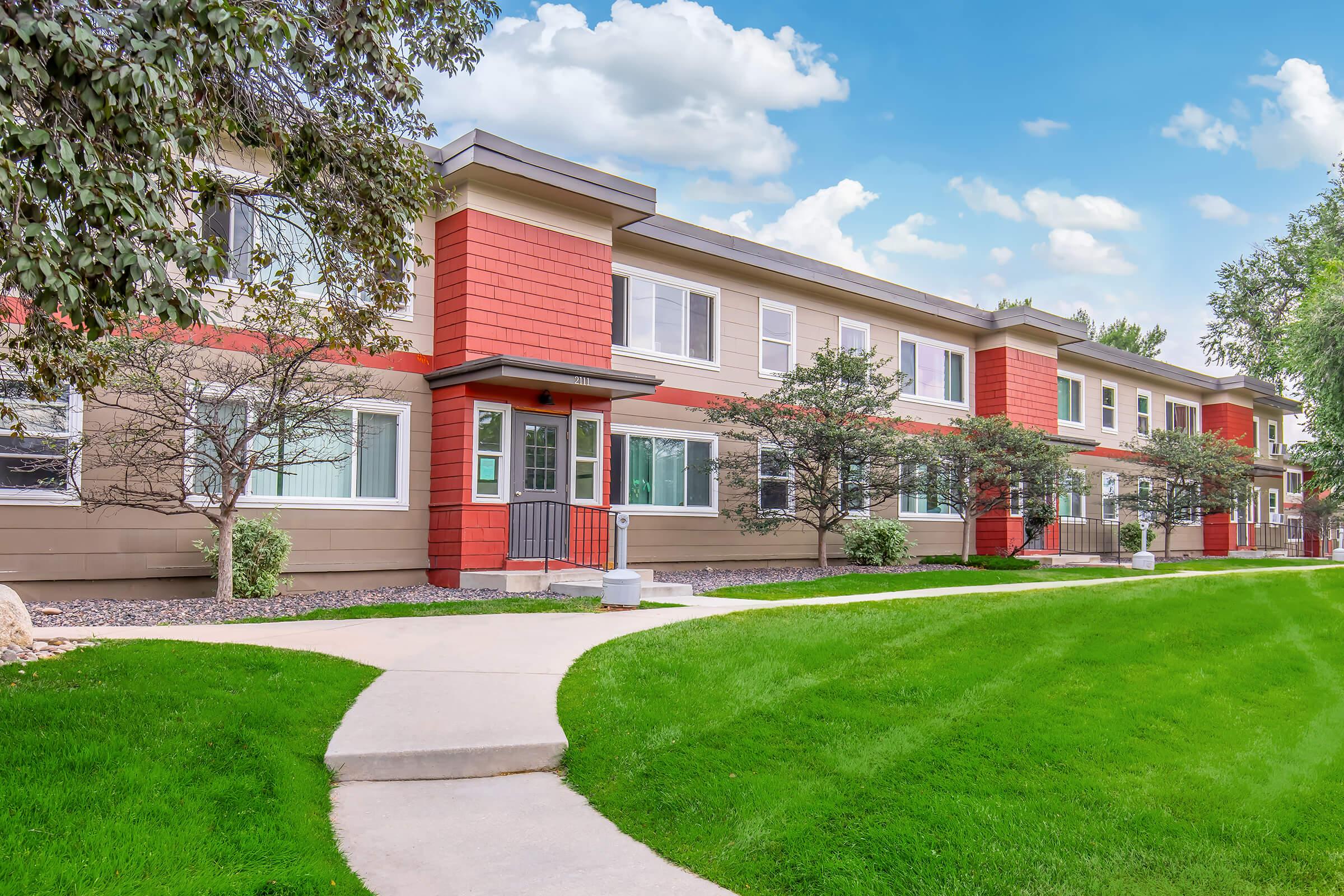 A row of modern, multi-unit apartments with colorful exterior featuring a blend of beige and red sections. Well-maintained landscaping includes green grass and small trees. A concrete walkway leads up to the entrance, under a blue sky with scattered clouds.