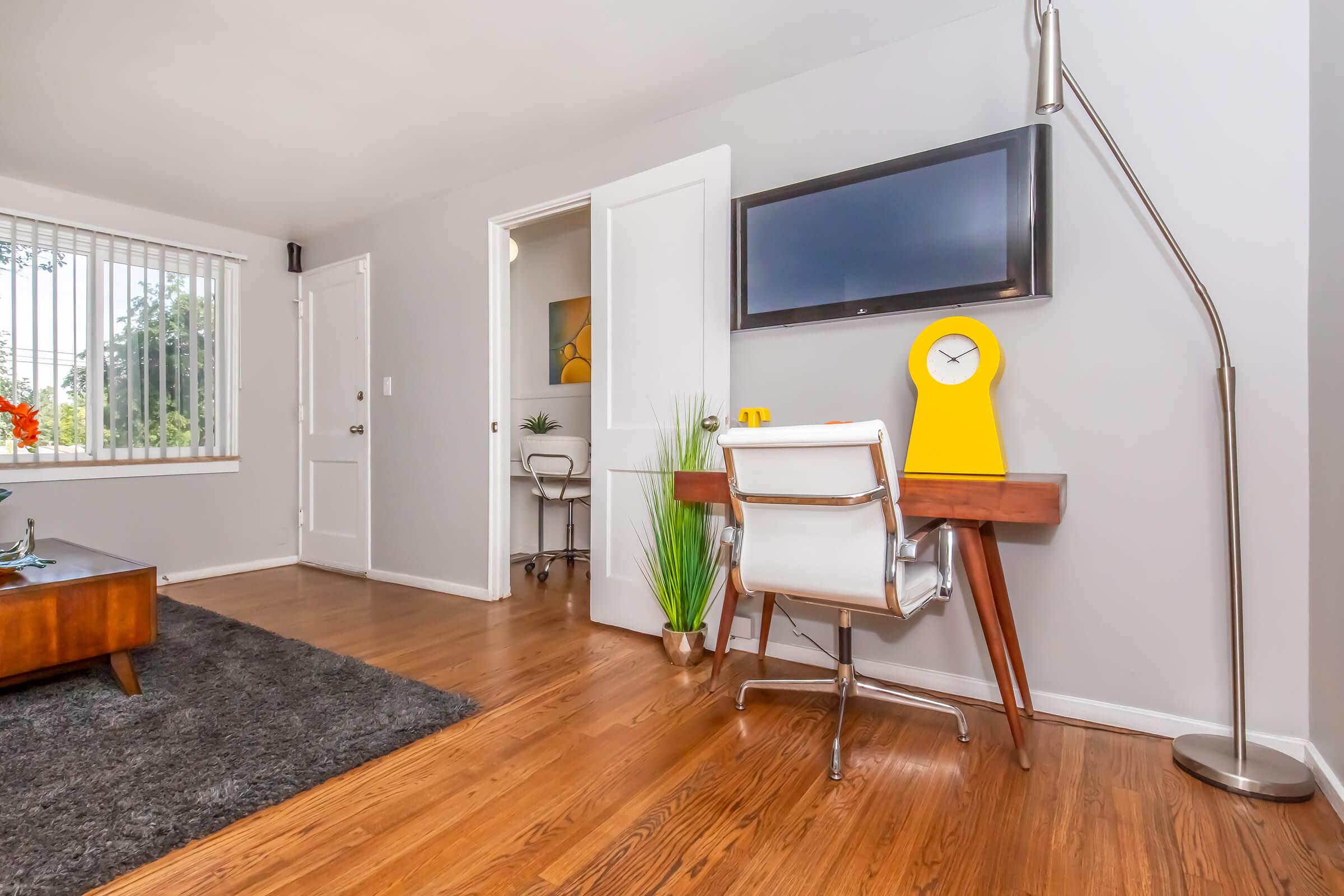A modern and stylish home office setup featuring a wooden desk with a yellow clock and decorative items, a comfortable white chair, a floor lamp, and a plush gray rug. The walls are painted in a light gray, with a large window letting in natural light, and a plant adding a touch of greenery to the space.