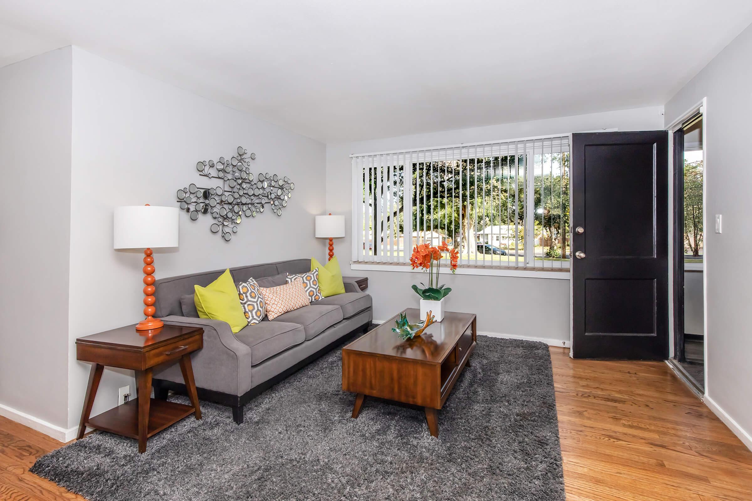 A cozy living room featuring a gray sofa with colorful yellow and patterned cushions, two lamps with orange bases, a wooden coffee table with an ornamental plant, and a textured gray area rug. Large windows allow for natural light, and there is a front door visible on the right side.