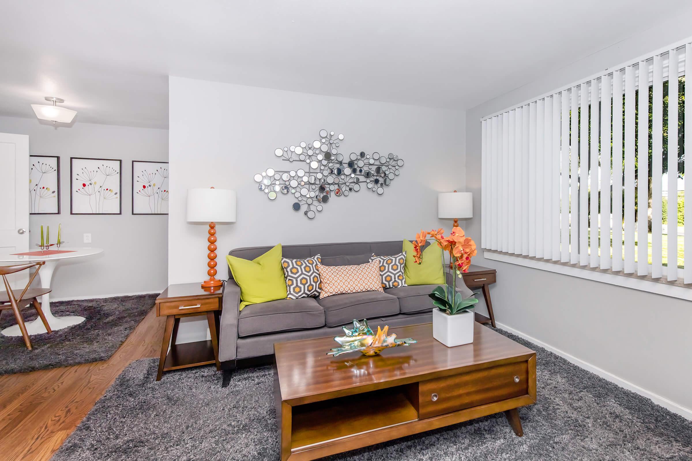 A modern living room featuring a gray sofa with colorful throw pillows, a wooden coffee table, and an orange flower arrangement. Two stylish lamps flank the sofa. The room has light gray walls, a plush dark rug, and a set of framed artwork on the wall. Natural light comes through vertical blinds.