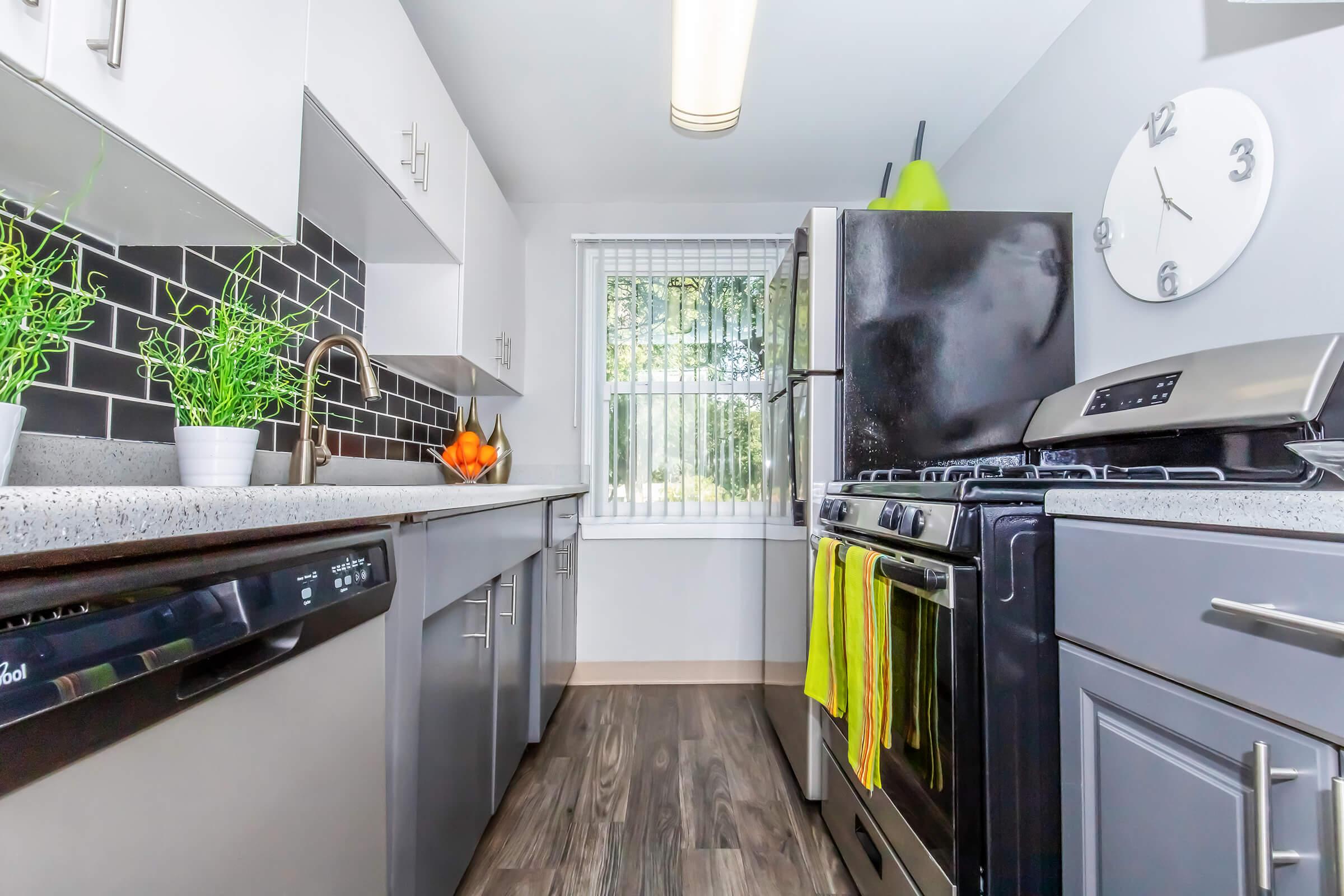 A modern kitchen featuring sleek gray cabinets, a black stove, and stainless steel appliances. The countertop is a light gray, with potted plants and a bowl of oranges for decoration. There's a window with sheer blinds, allowing natural light to enter, and a clock on the wall. Brightly colored kitchen towels hang from the stove.