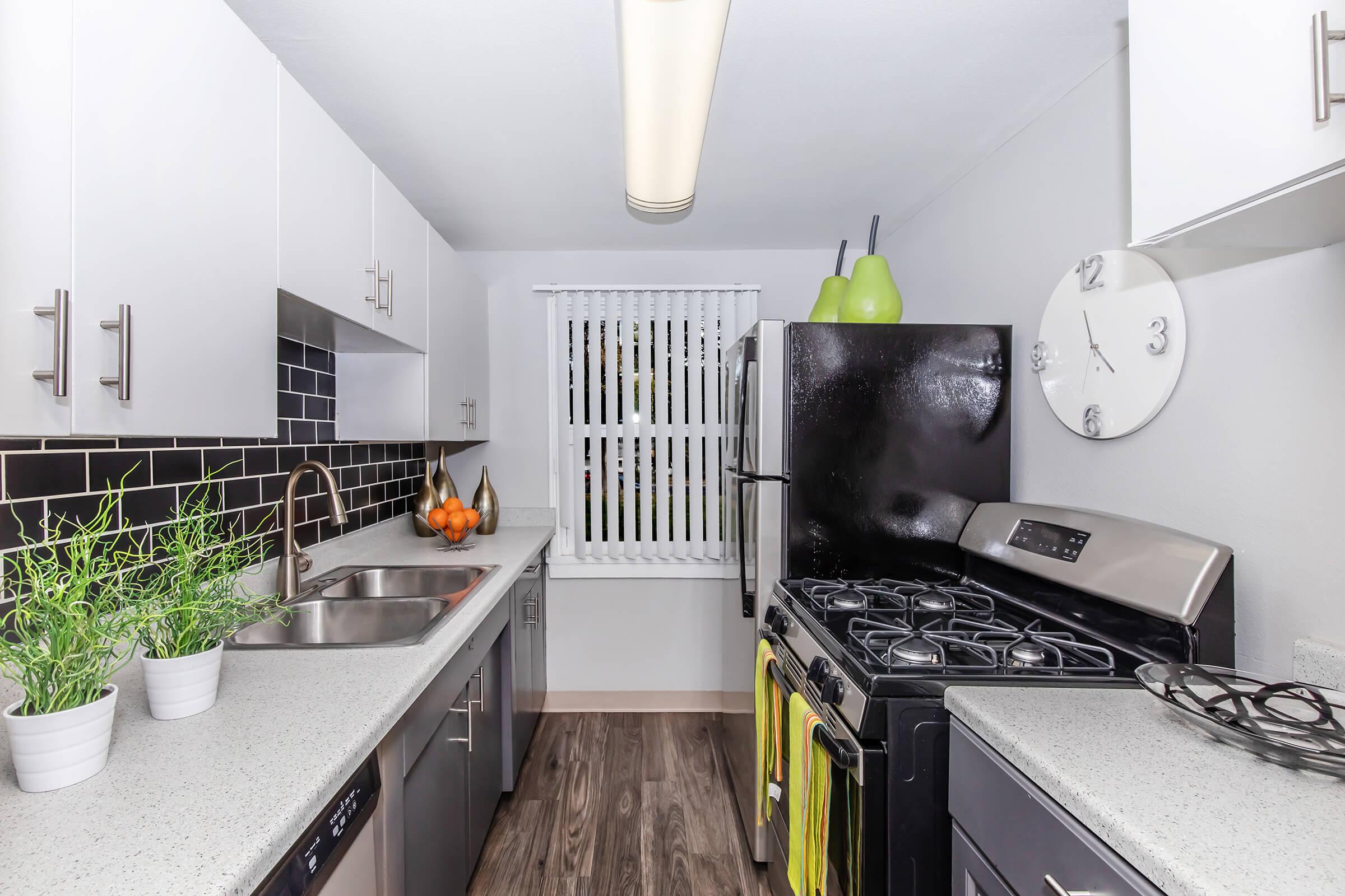A modern kitchen featuring white cabinetry, a black refrigerator, and a stainless steel gas stove. The countertop is light gray with a double sink, and there are two potted plants on the counter. A wall clock and decorative fruits in a bowl add color, while a window with blinds allows natural light to enter.
