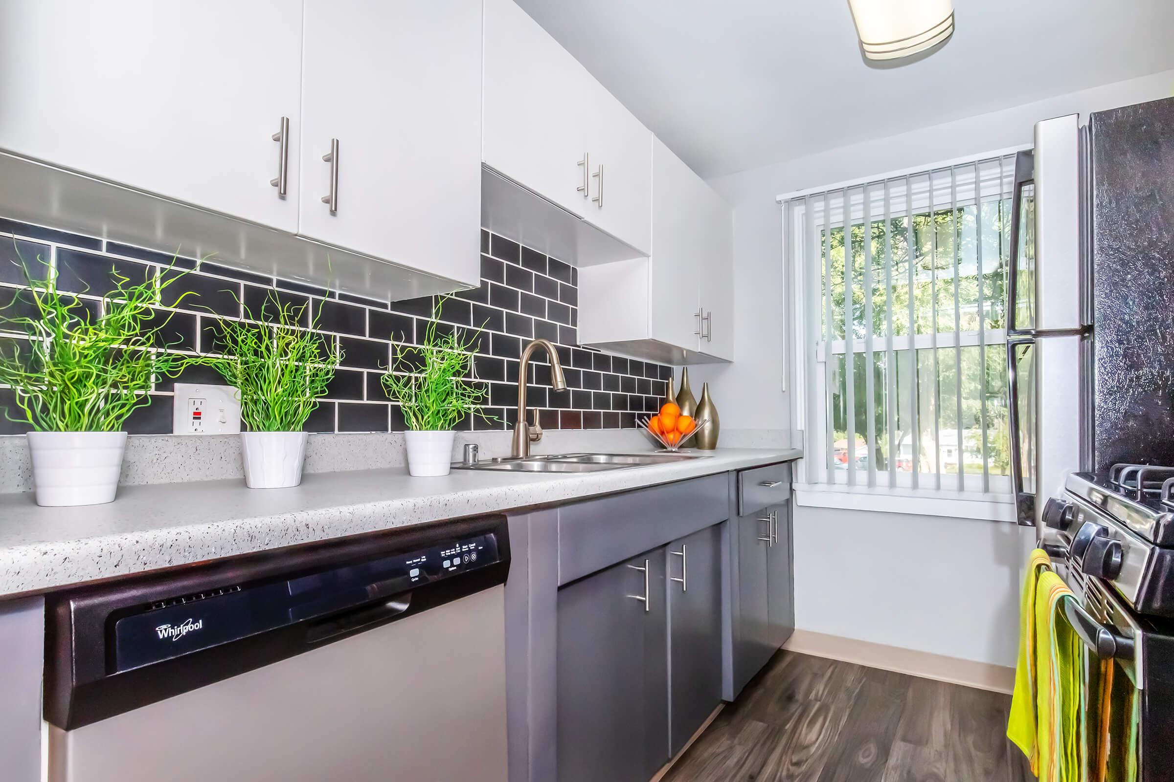 Modern kitchen featuring sleek gray cabinets, a stainless steel sink, and a black tiled backsplash. Brightly lit by a window with white blinds, the kitchen includes potted green plants, a bowl of oranges, and appliances like a dishwasher and stove. A vibrant yellow dish towel hangs from the stove.