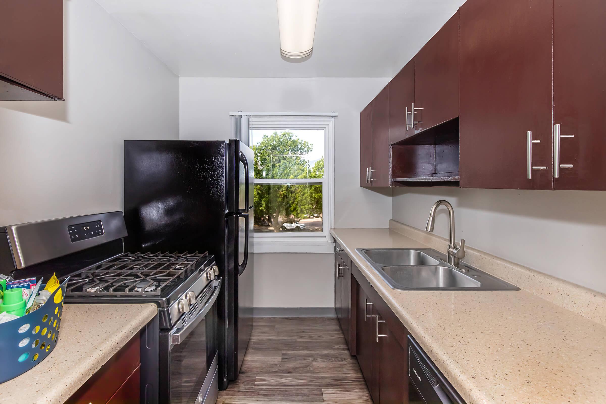 A modern kitchen featuring dark wooden cabinets, a stainless steel oven and gas stove, a black refrigerator, and a double sink with a chrome faucet. The countertop is a light beige material. There's a window allowing natural light, with trees visible outside. The flooring is a wood-like finish.