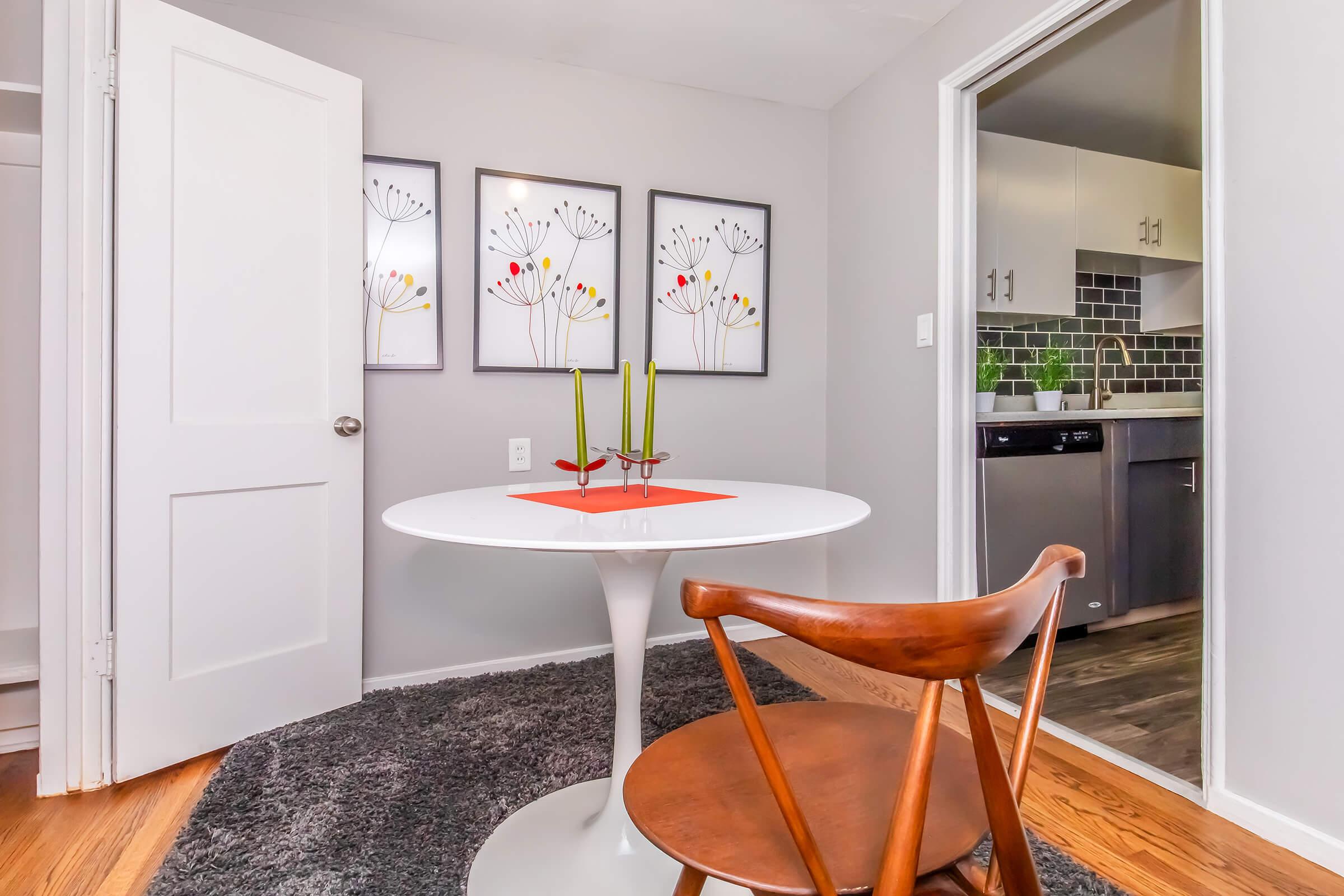A modern dining area featuring a round white table with an orange centerpiece and two green candles. Behind the table, there are three framed floral prints on the gray wall. A wooden chair sits next to the table on a plush gray rug. A doorway leads to a kitchen with dark cabinetry in the background.