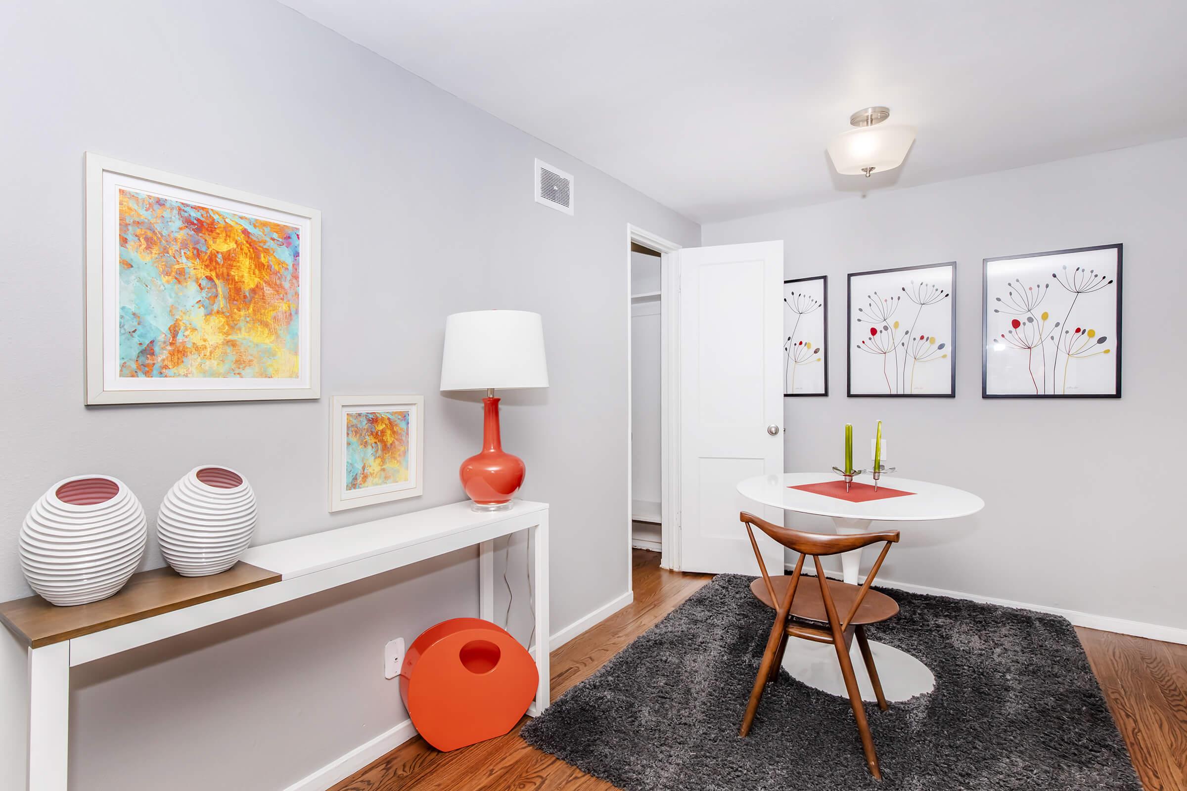 A modern dining area featuring a small round table with a white top and wooden legs, accompanied by a wooden chair. The walls are decorated with colorful abstract art and floral prints. A sleek white console table holds two white vases, and a stylish lamp, while a red accent piece adds a pop of color to the space. A soft gray rug completes the look.