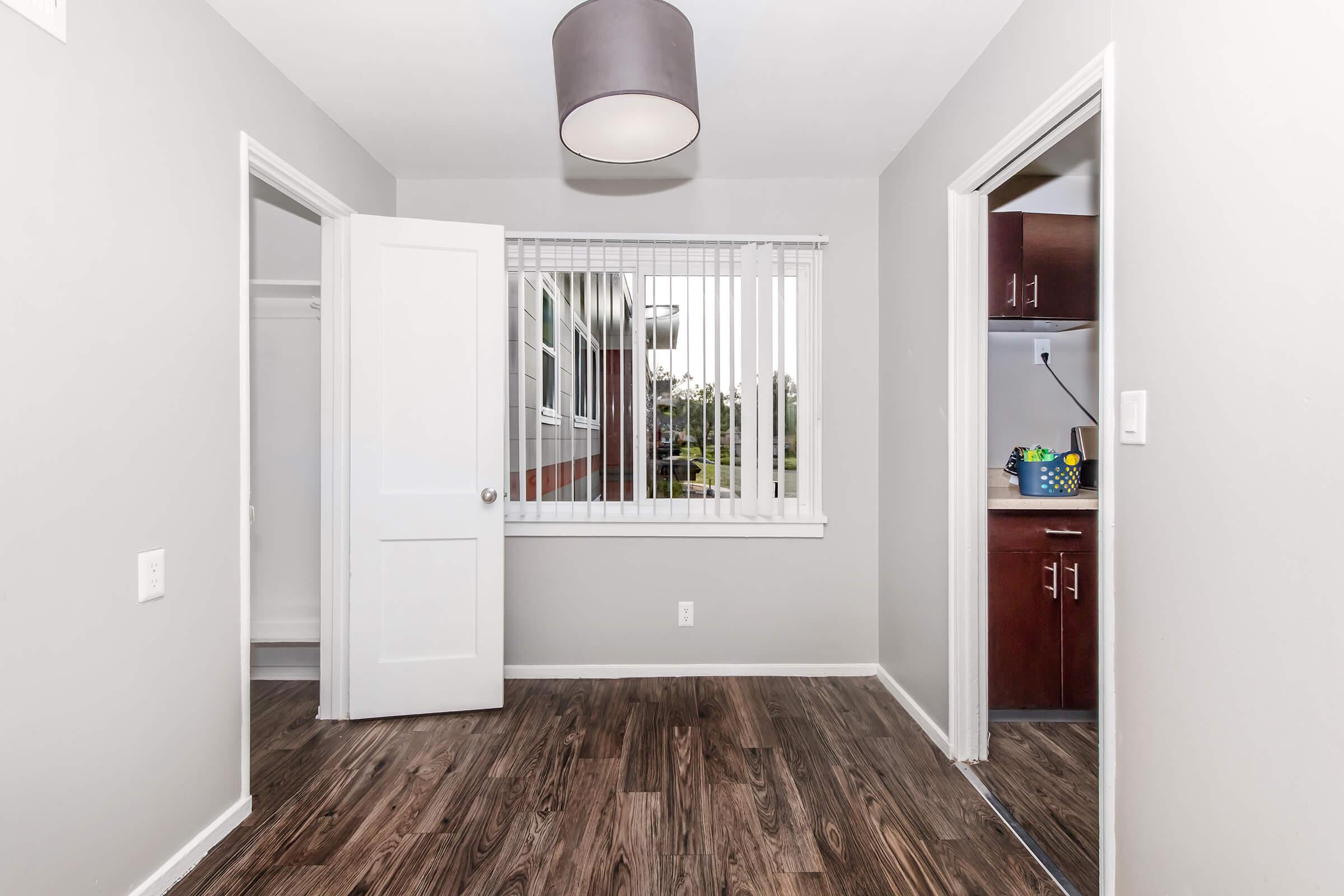 A well-lit, neutral-colored room featuring a light fixture, a window with blinds, and two doorways leading to adjacent spaces. The flooring is dark wood, and the room has a simple, modern design with no furniture.