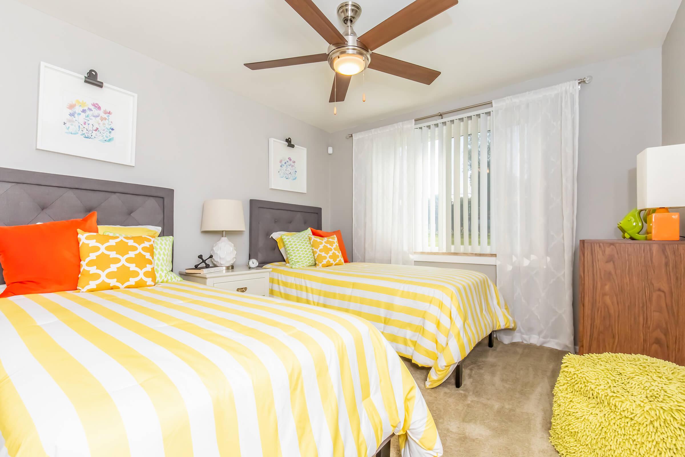 A bright and modern bedroom featuring two beds with yellow and white striped bedding. One bed has a decorative pillow. A bedside lamp and artwork are on the walls, and a large window with sheer curtains allows natural light to enter. The room has a cozy and inviting atmosphere.