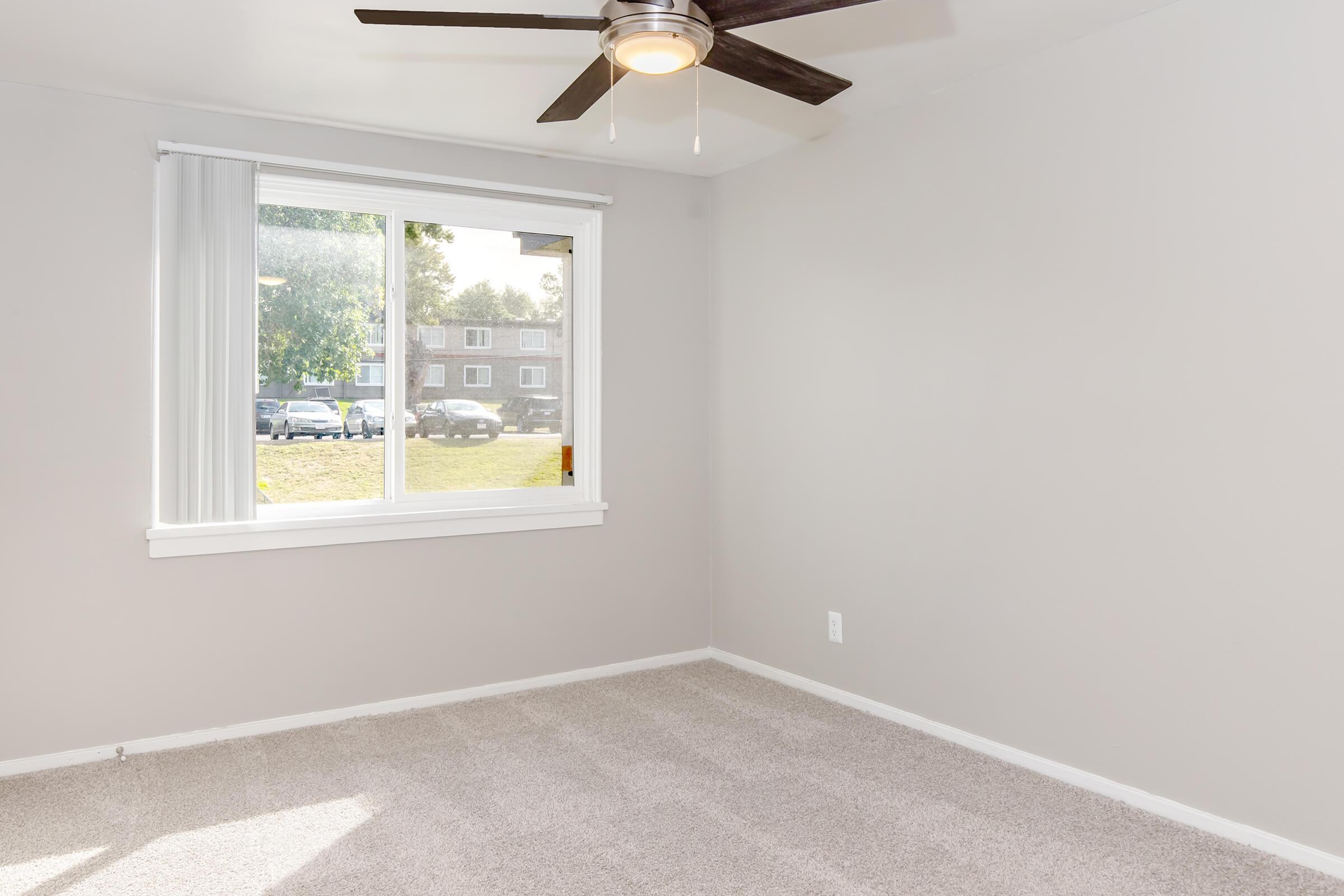 A small, empty room with light gray walls and a ceiling fan. There is a large window that allows natural light to enter, showing a view of a grassy area and parked cars outside. The floor is covered with light-colored carpet.