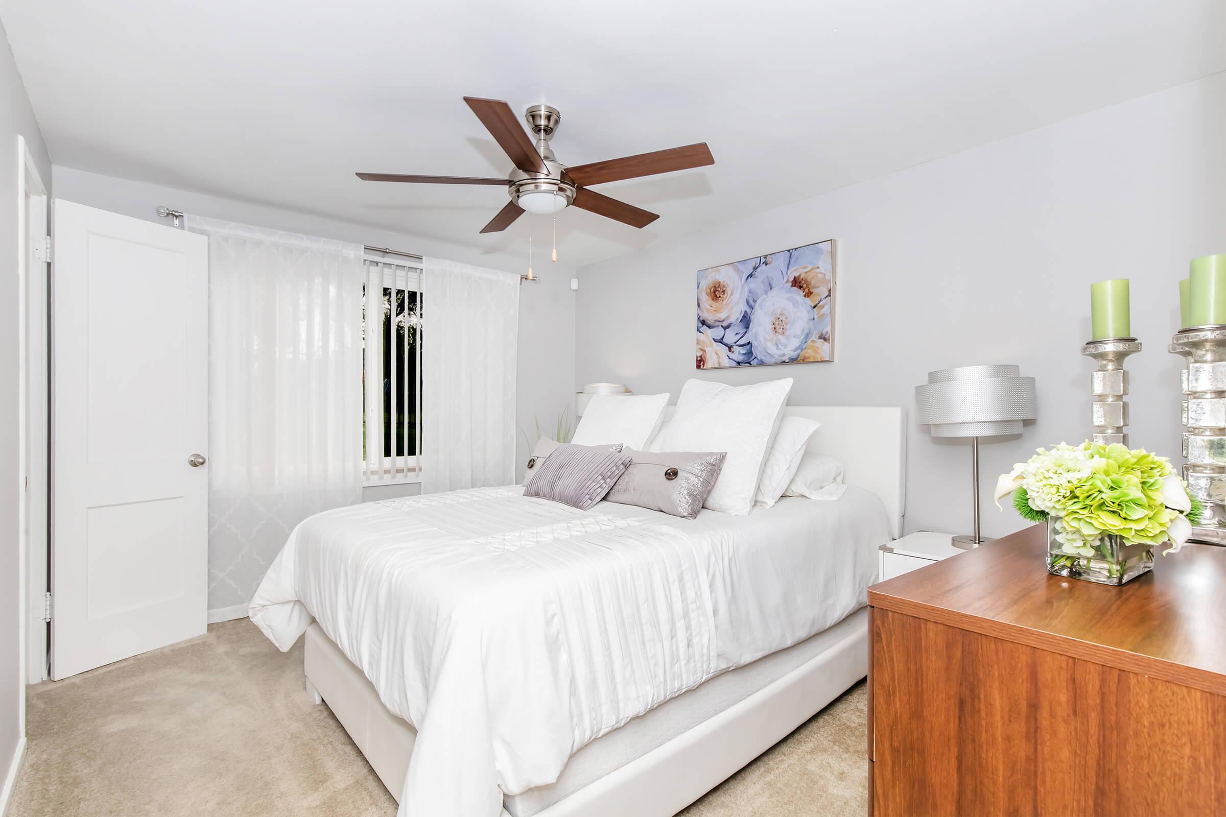 A cozy bedroom featuring a white bed with decorative pillows, a ceiling fan, and soft lighting. There's a wooden nightstand with decorative green flowers and candles. A piece of floral artwork adorns the wall, and sheer white curtains allow natural light to enter the space. The floor is carpeted for a warm feel.