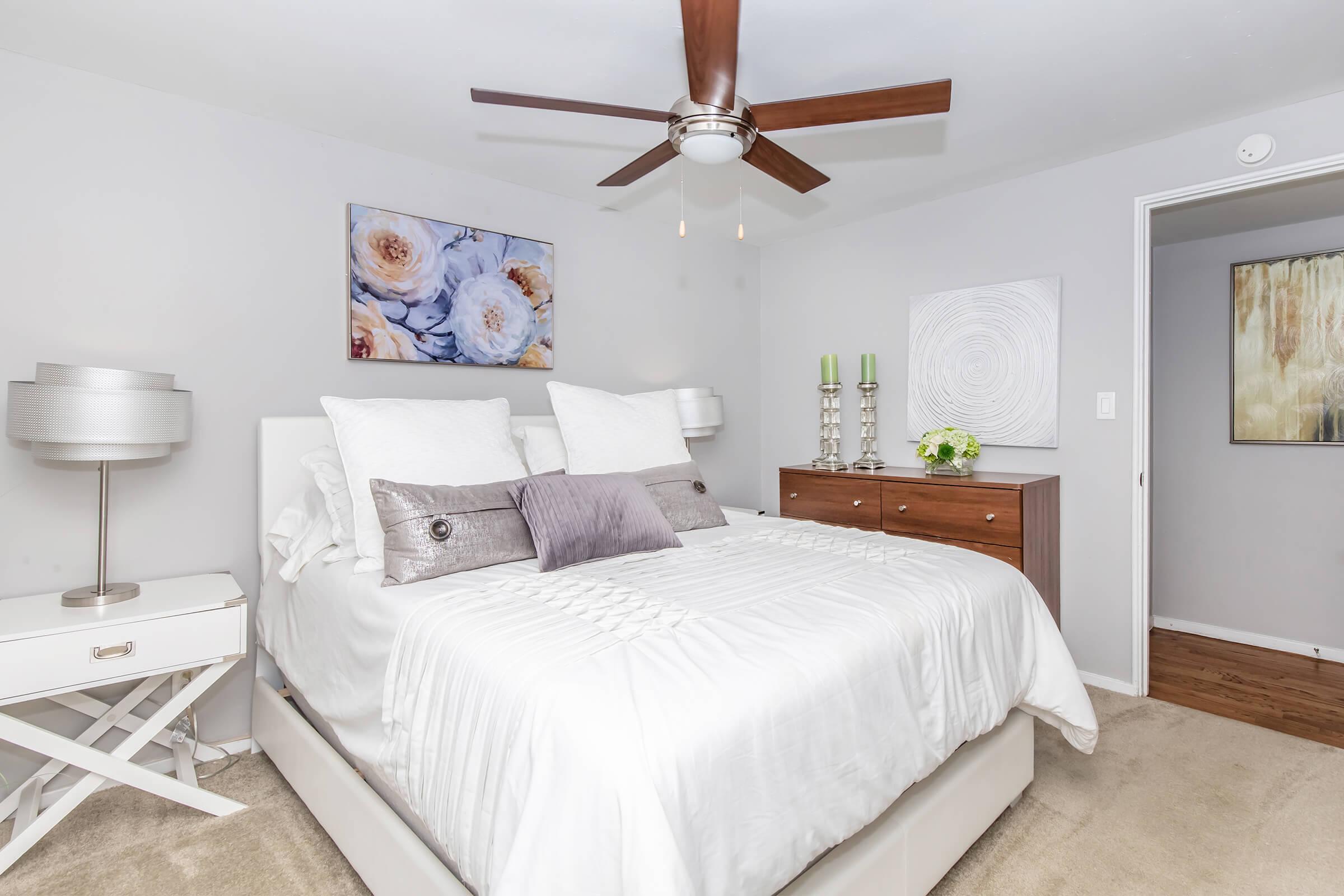 A cozy bedroom featuring a queen-sized bed with white bedding and decorative pillows. There are two bedside lamps, a wooden dresser with decorative items, and wall art depicting flowers and abstract designs. A ceiling fan enhances the airy feel and light gray walls add a calm ambiance.
