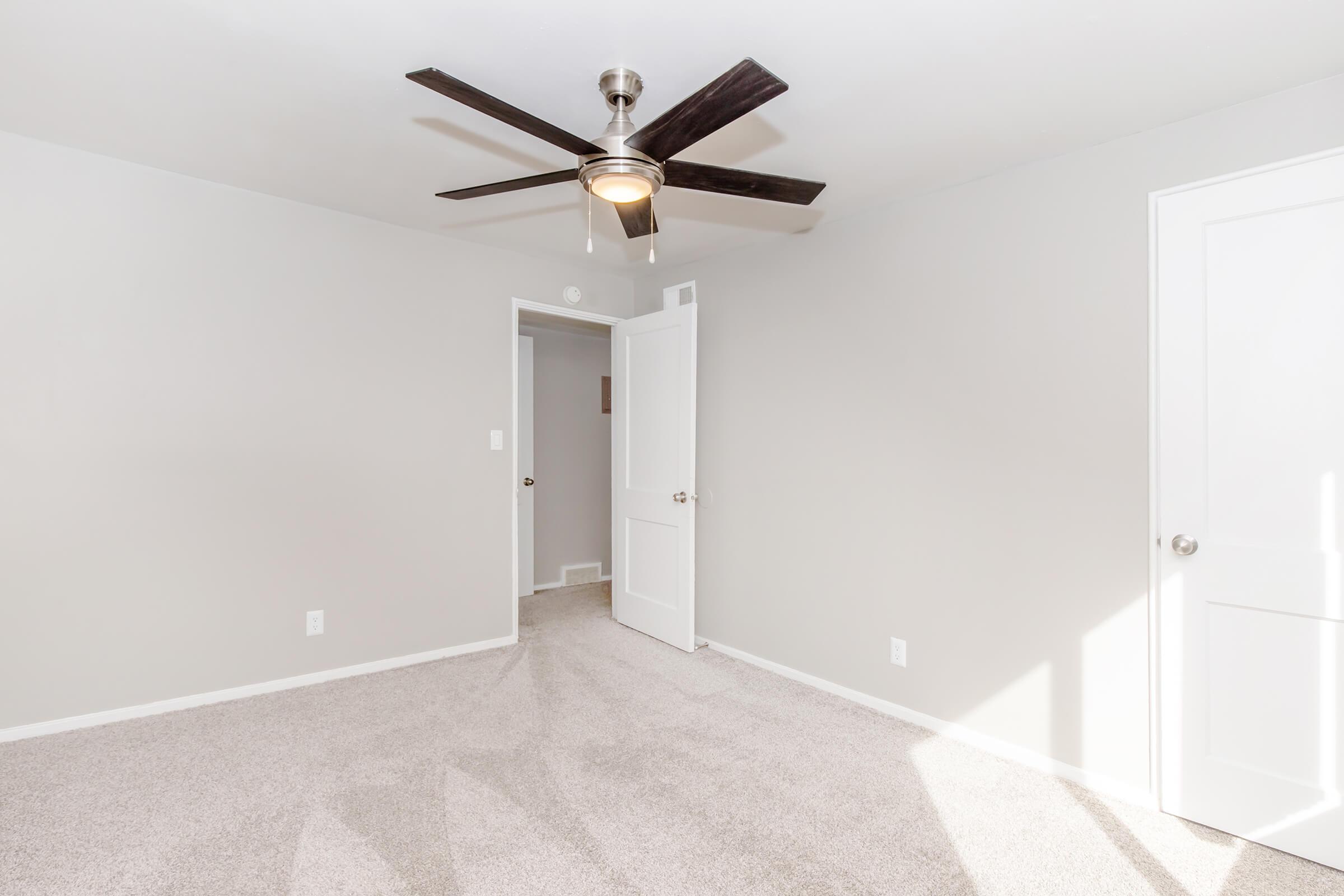A spacious, empty room with light gray walls and a plush carpet. A ceiling fan with five blades hangs from the ceiling, and there are two white doors leading to other areas. Natural light enters the room, creating a bright, inviting atmosphere.