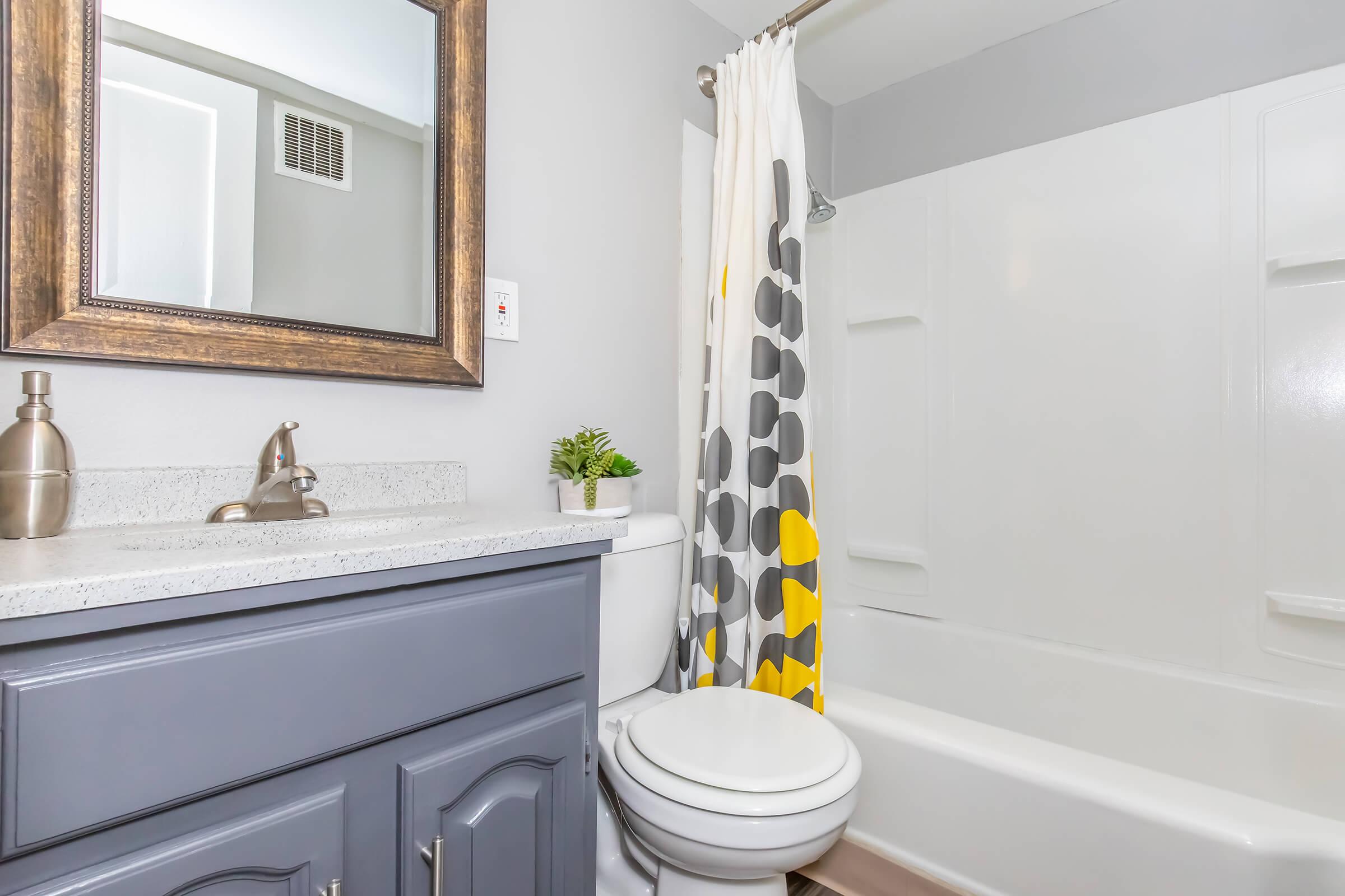A clean, modern bathroom featuring a gray and white color scheme. There is a sleek countertop with a decorative vase, a large mirror with a decorative frame, a bathtub with a shower curtain displaying a geometric pattern in yellow and gray, and a potted plant for a touch of greenery.
