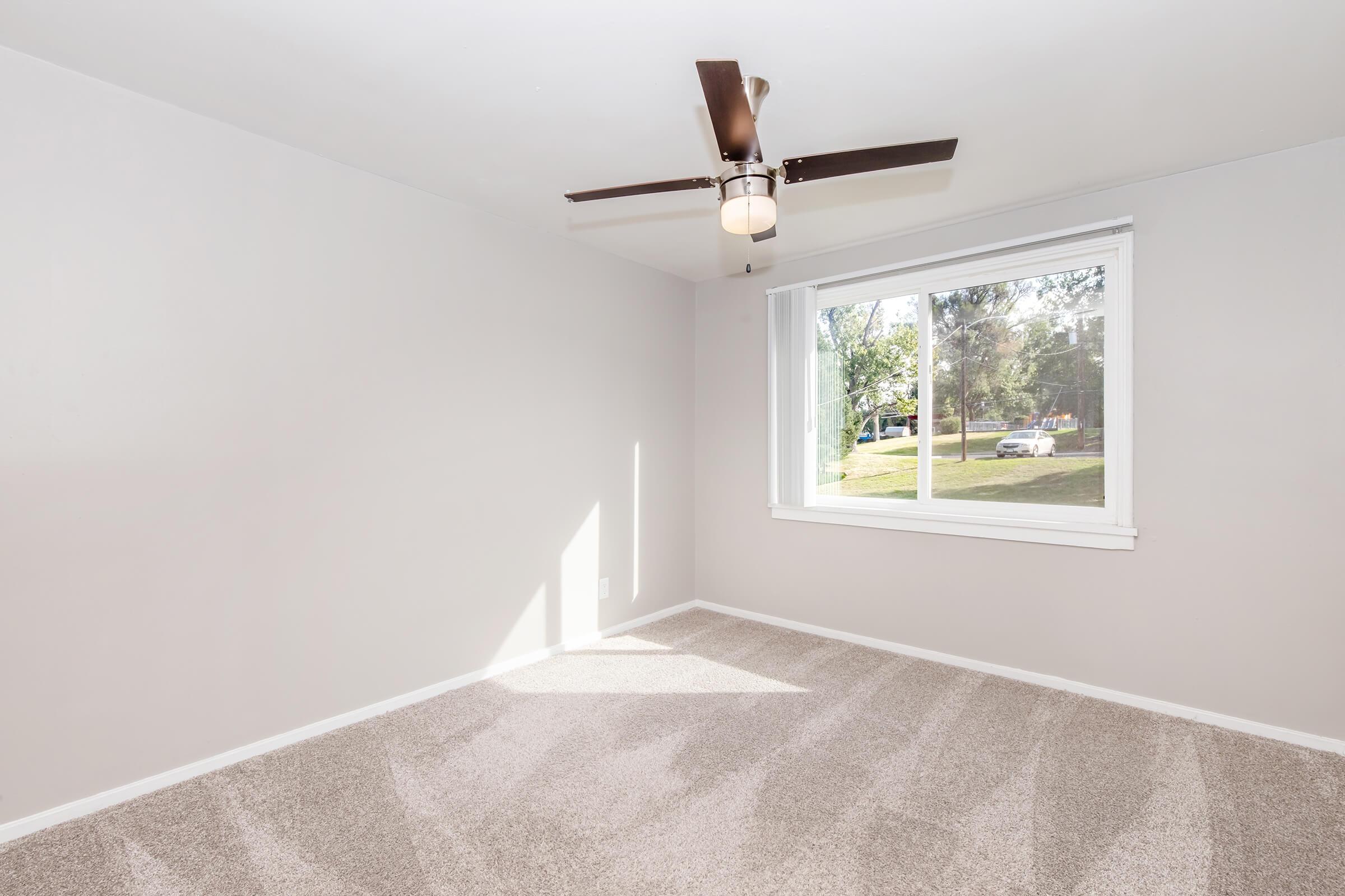 Empty room with a freshly painted light gray wall, a ceiling fan with light fixture, and large window allowing natural light. The floor is covered in soft, beige carpet. Outside the window, greenery and cars are visible. The room appears spacious and inviting.